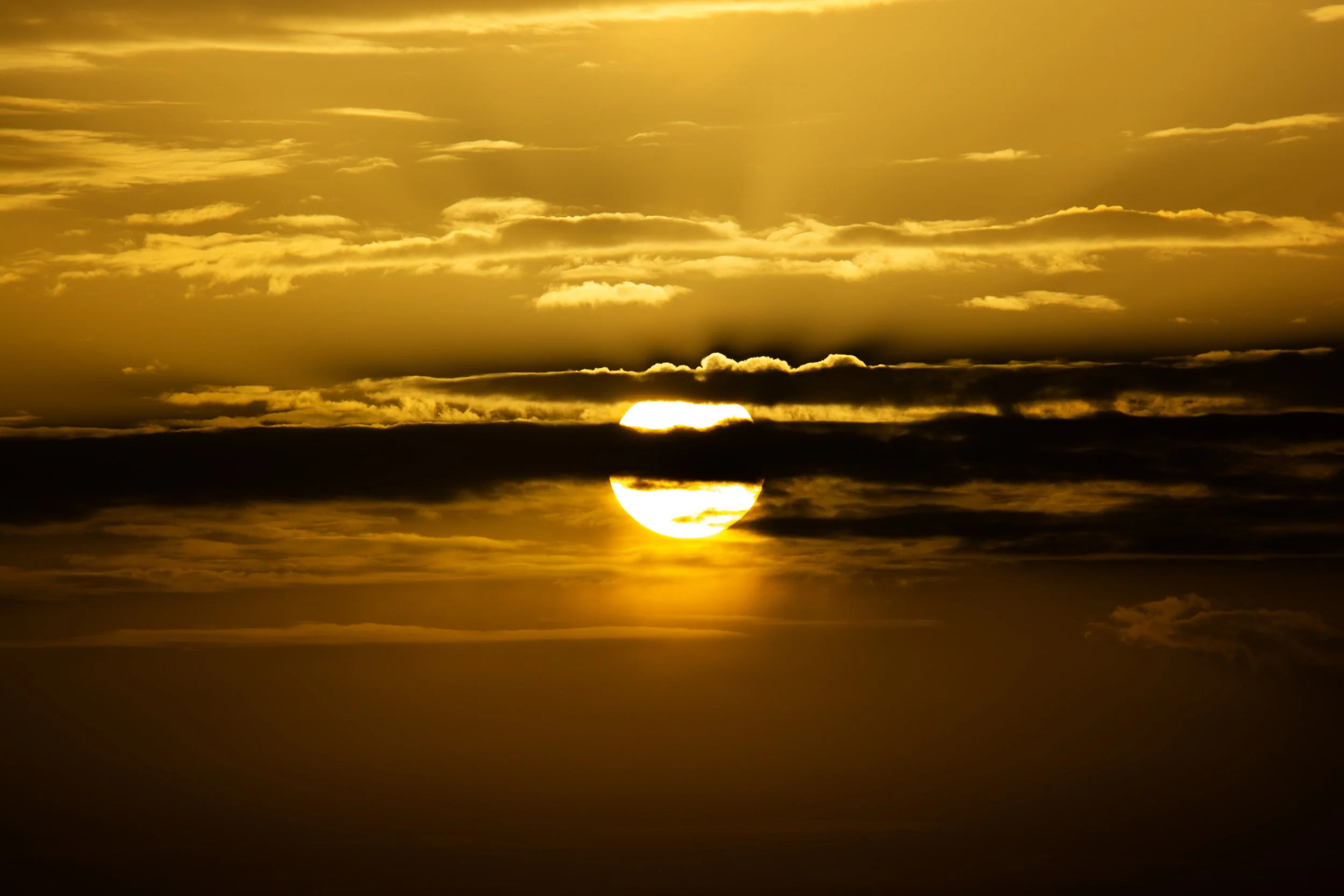 Sun setting behind clouds, creating a golden sky with dark silhouettes of clouds.
