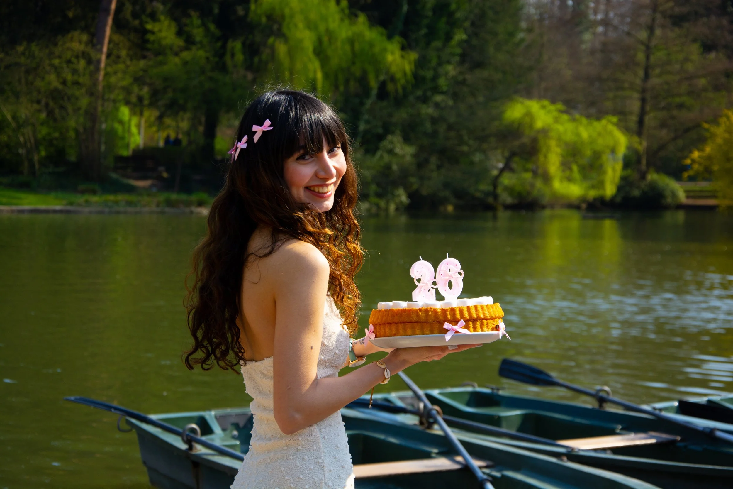 A young woman in a white dress holding a birthday cake with candles, standing near a lake with boats and trees in the background, celebrating her 26th birthday.