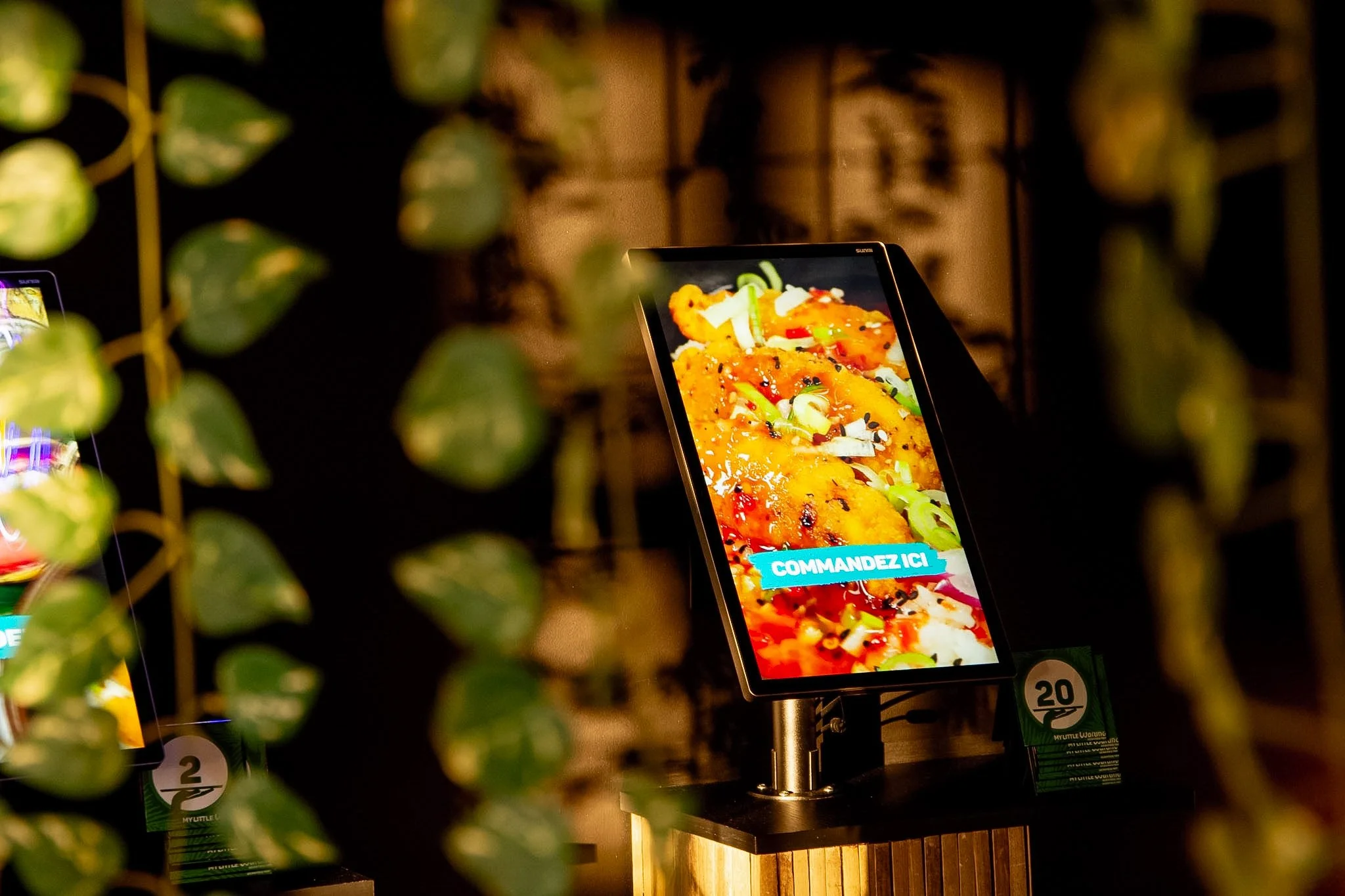 Digital menu screen with colorful food image and blue text 'COMMANDEZ ICI' in a restaurant setting, surrounded by green leaves in the foreground.