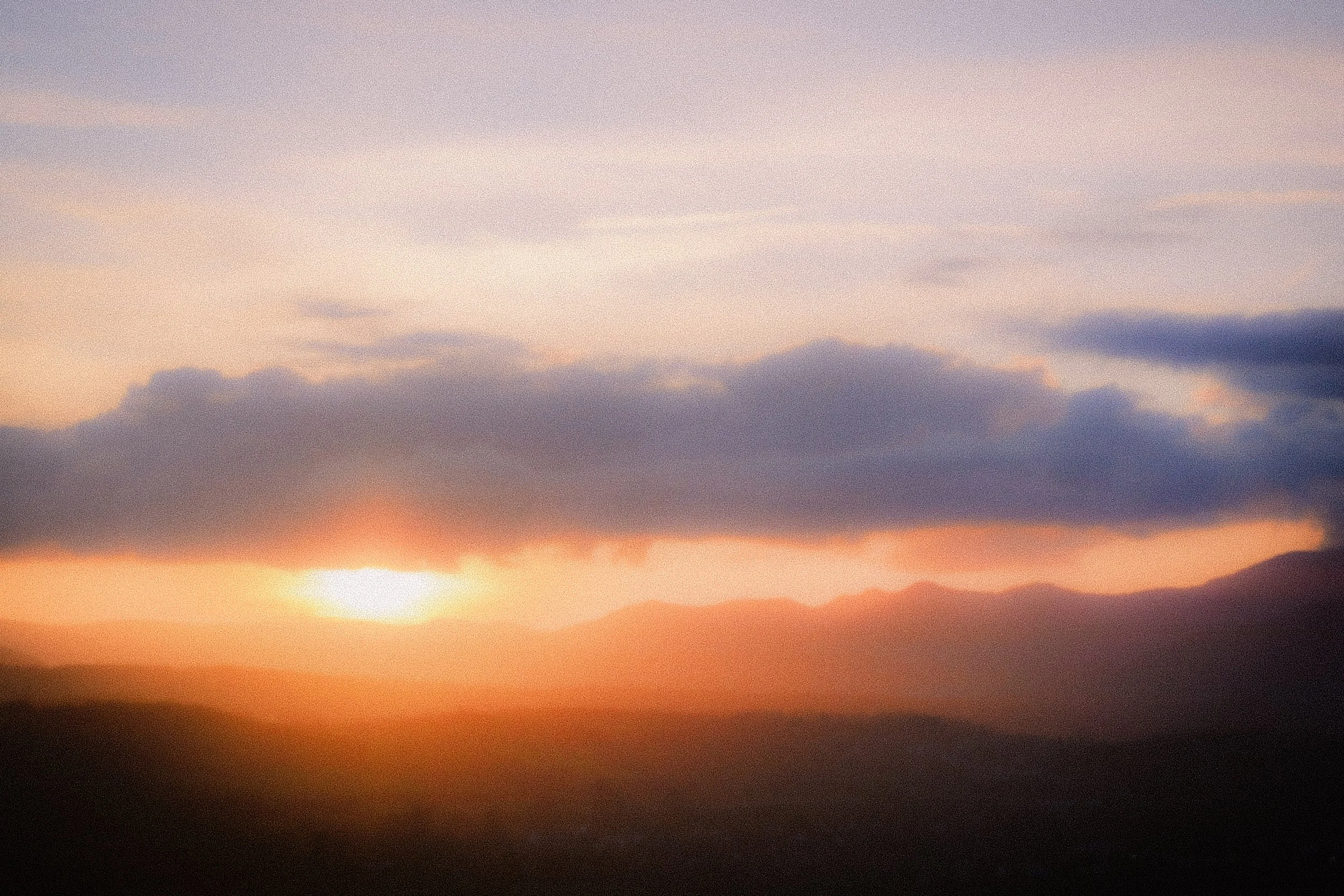 A sunset over a mountainous landscape with a sky filled with clouds, displaying shades of orange, purple, and blue.