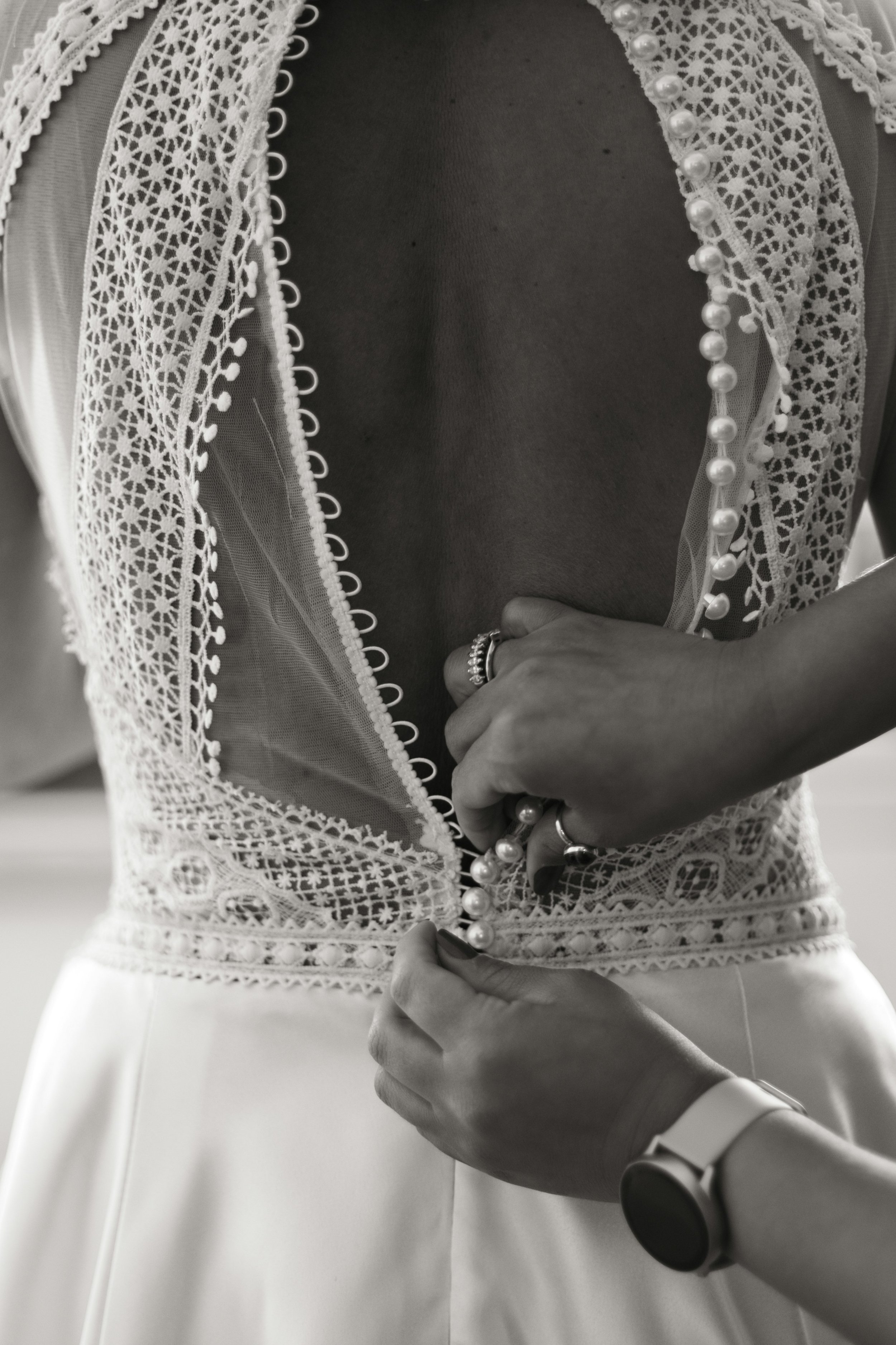 Nahaufnahme eines Schmuckstücks bei der Anprobe an einer Frau, die ein weißes Kleid trägt. Es handelt sich um eine enganliegende, elegante Kleid mit aufwändiger Lochstickerei und Perlenstickerei am Ausschnitt, das vermutlich für eine besondere Gelegenheit getragen wird.