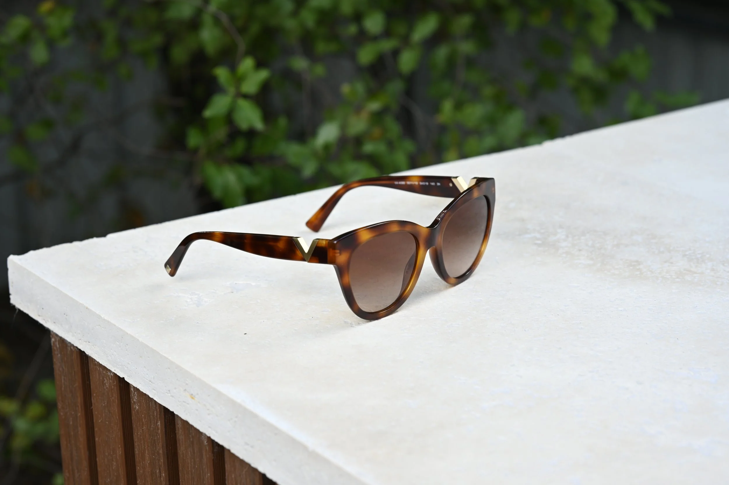 A pair of tortoise shell sunglasses resting on natural travertine surface with a background of green foliage.