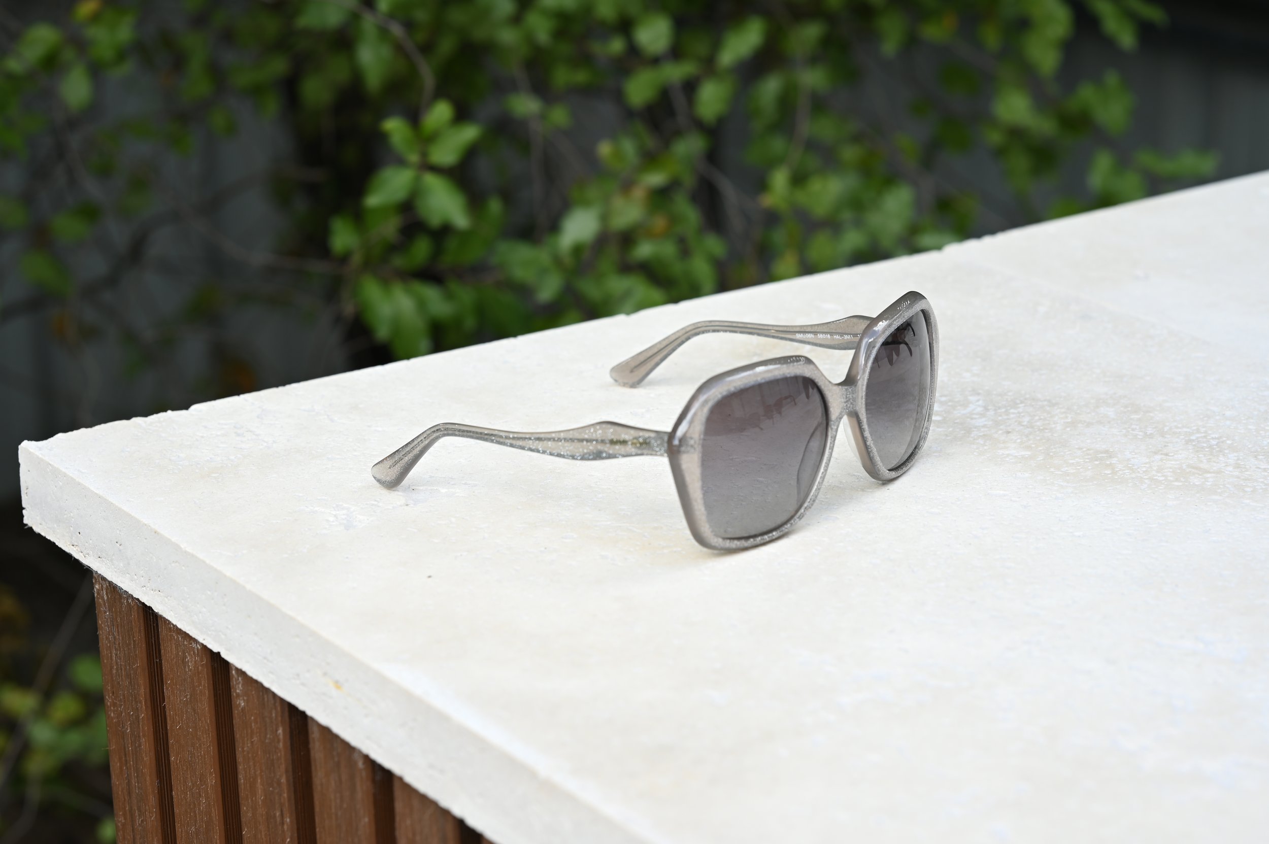 A pair of grey Miu Miu sunglasses resting on natural stone surface outdoors with a  green leafy background.