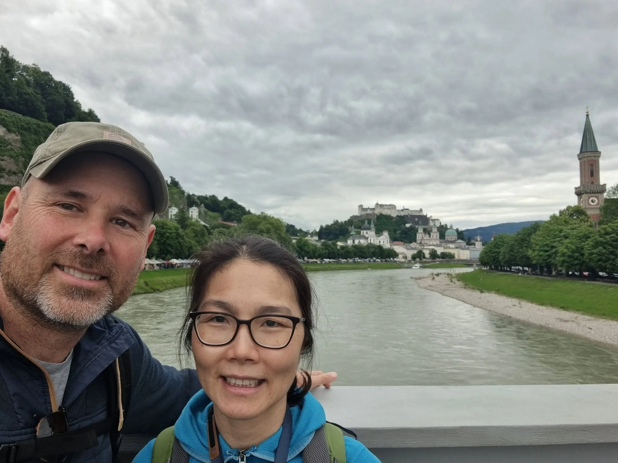 A man and a woman pose for a selfie near a river with a historic European town and castle on a hill in the background. The man is wearing a cap and jacket, and the woman has glasses and a backpack. The sky is overcast.