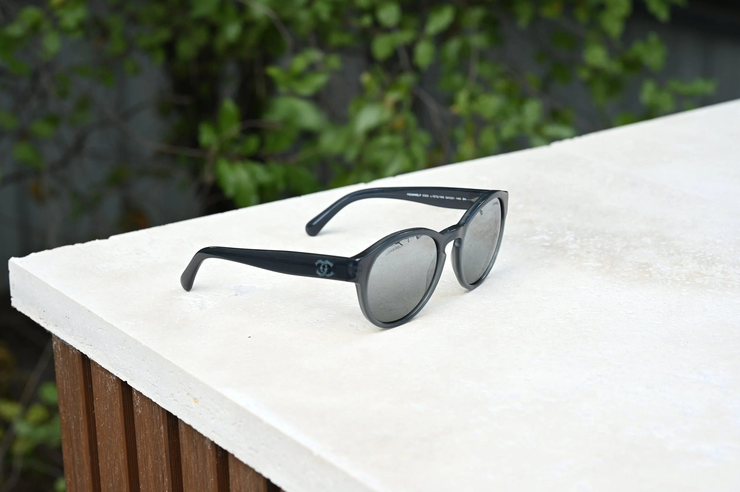 Pair of black Chanel sunglasses resting on a natural stone surface outdoors with greenery in the background.