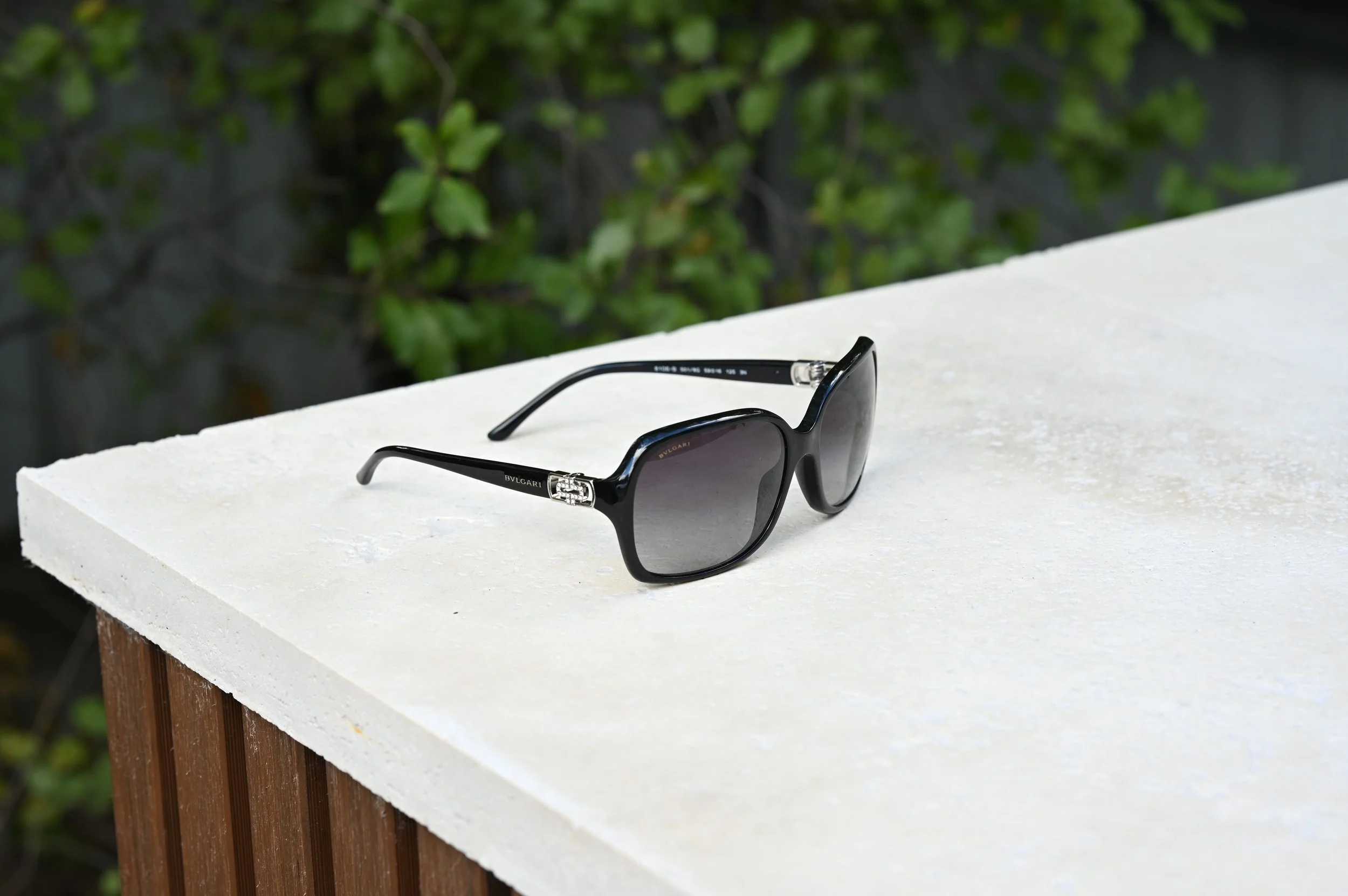 Black Bvlgari sunglasses with dark lenses and metal accents on the temples resting on a white textured travertine stone outdoors, with green foliage in the background.