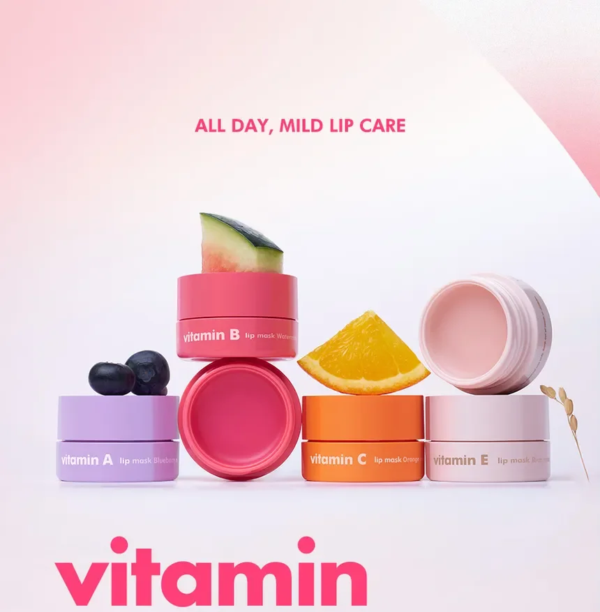 Colorful jars of lip mask ingredients labeled with vitamins A, B, C, and E, with slices of blueberry, watermelon, orange, and what appears to be grapefruit or other citrus, and a pink balm inside an open container. The background is light with pink accents, and the text reads 'All day, mild lip care' and 'vitamin'.
