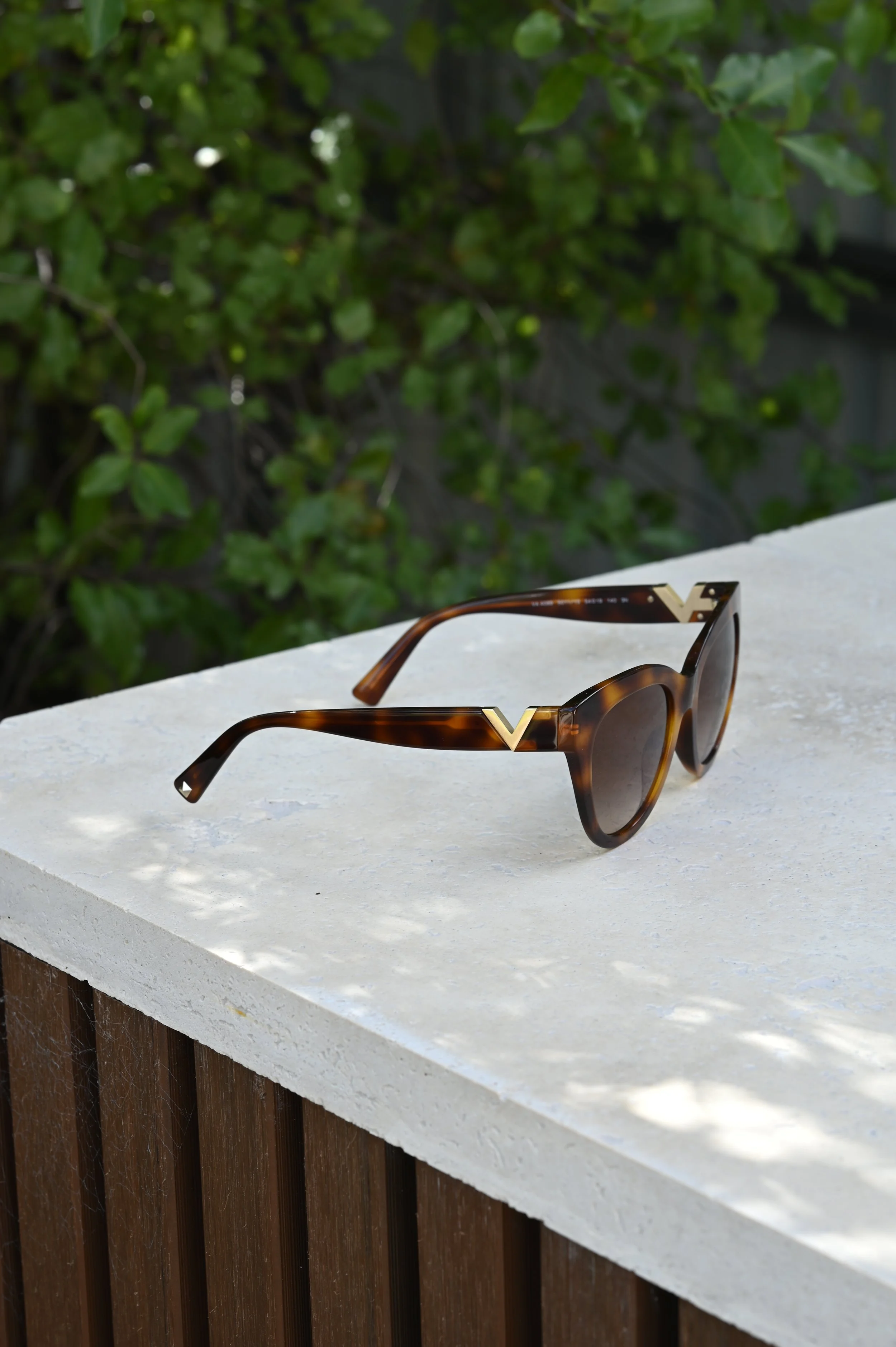 A pair of tortoise shell Valentino sunglasses resting on a natural stone outdoors, with green foliage in the background.
