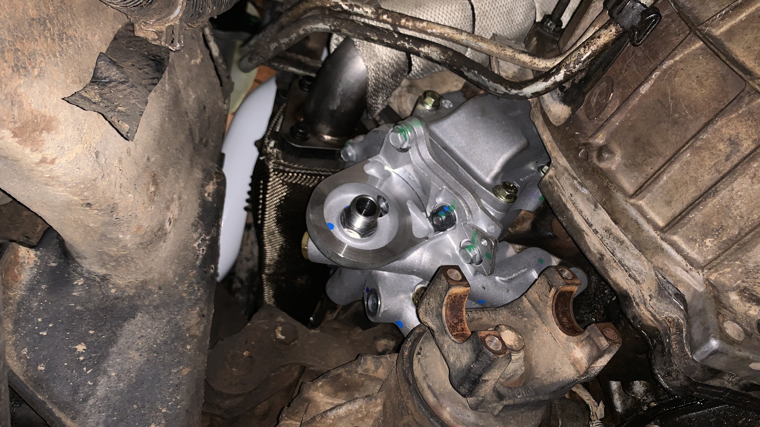 Close-up view of a new silver mechanical part installed within an engine, more rusted and dirty components surrounding it.