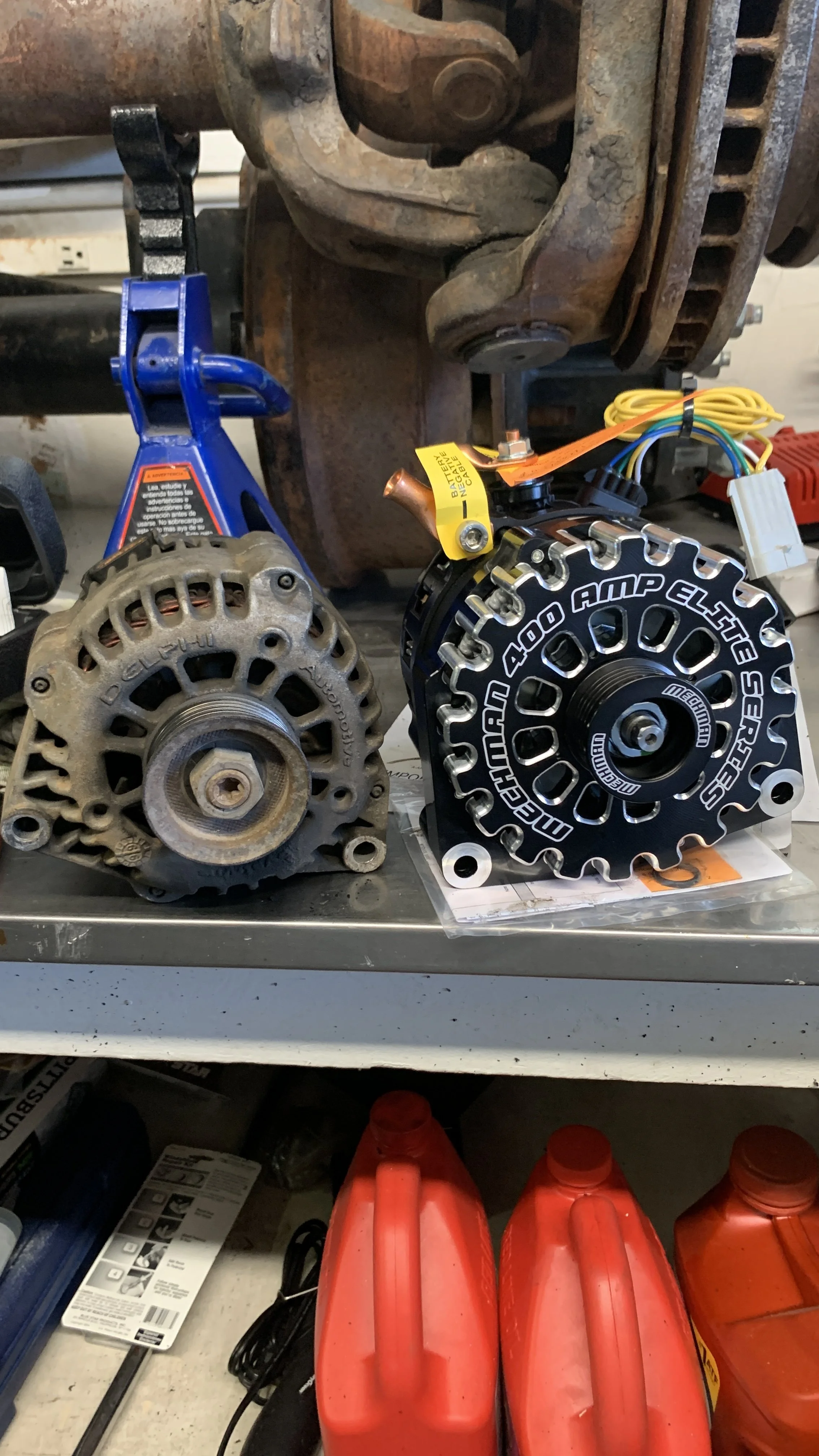 A car alternator and a high-performance electric motor placed on a workbench, with various tools and containers of automotive fluids in the background.