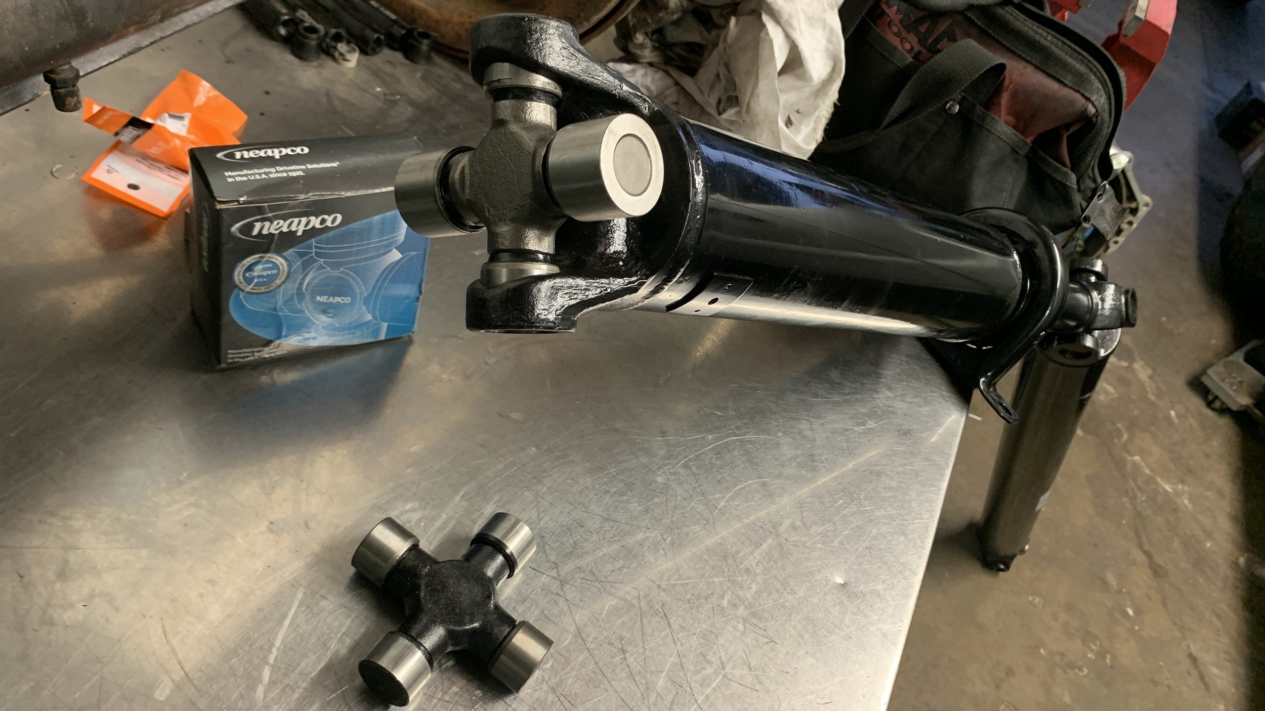 A black motorcycle front suspension fork tube, a wheel hub, and a box of Neapco brand parts on a metal workbench in a garage or workshop.