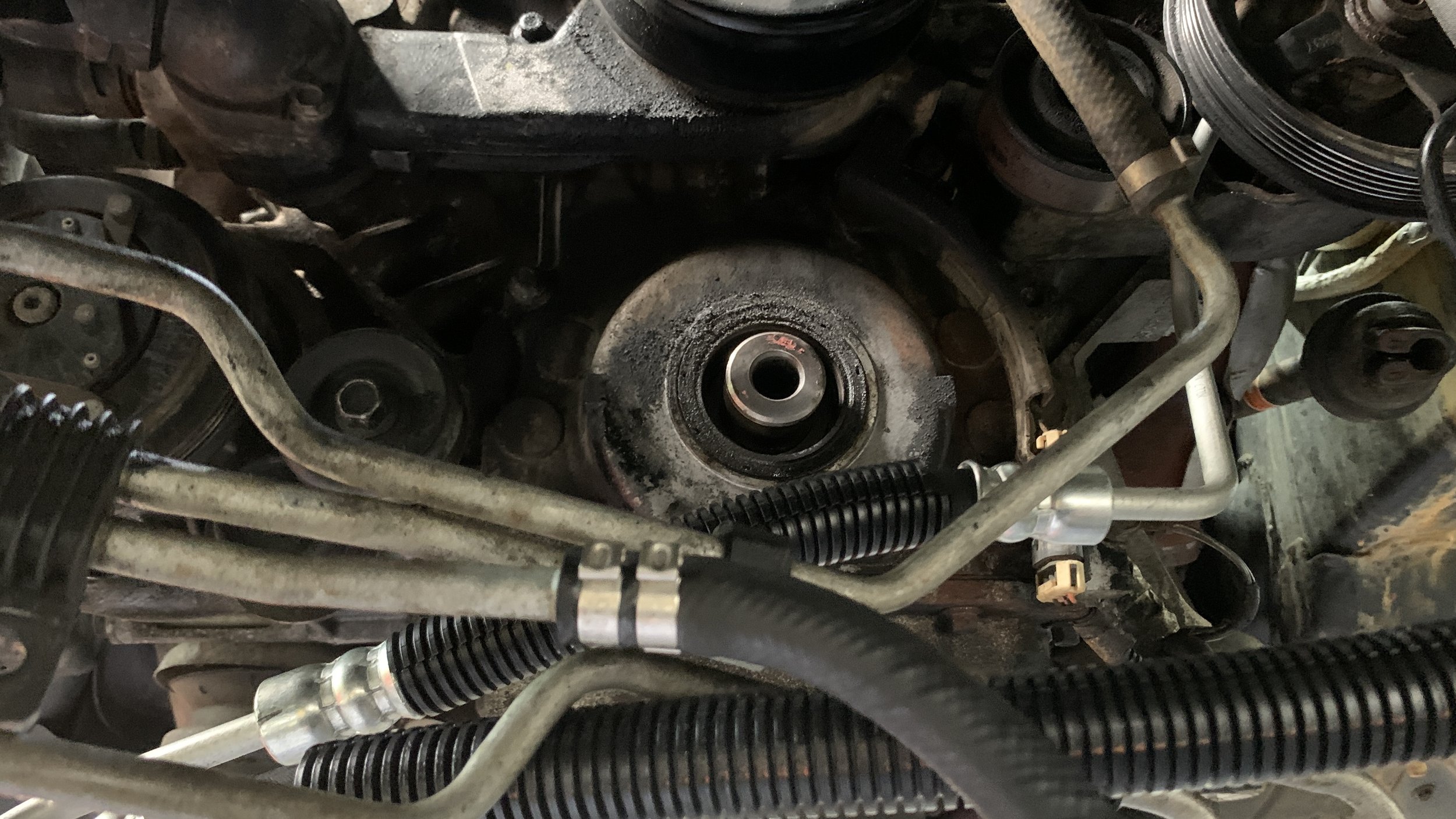 Close-up view of a car engine showing parts such as belts, pulleys, hoses, and metal components, with signs of dirt and wear.
