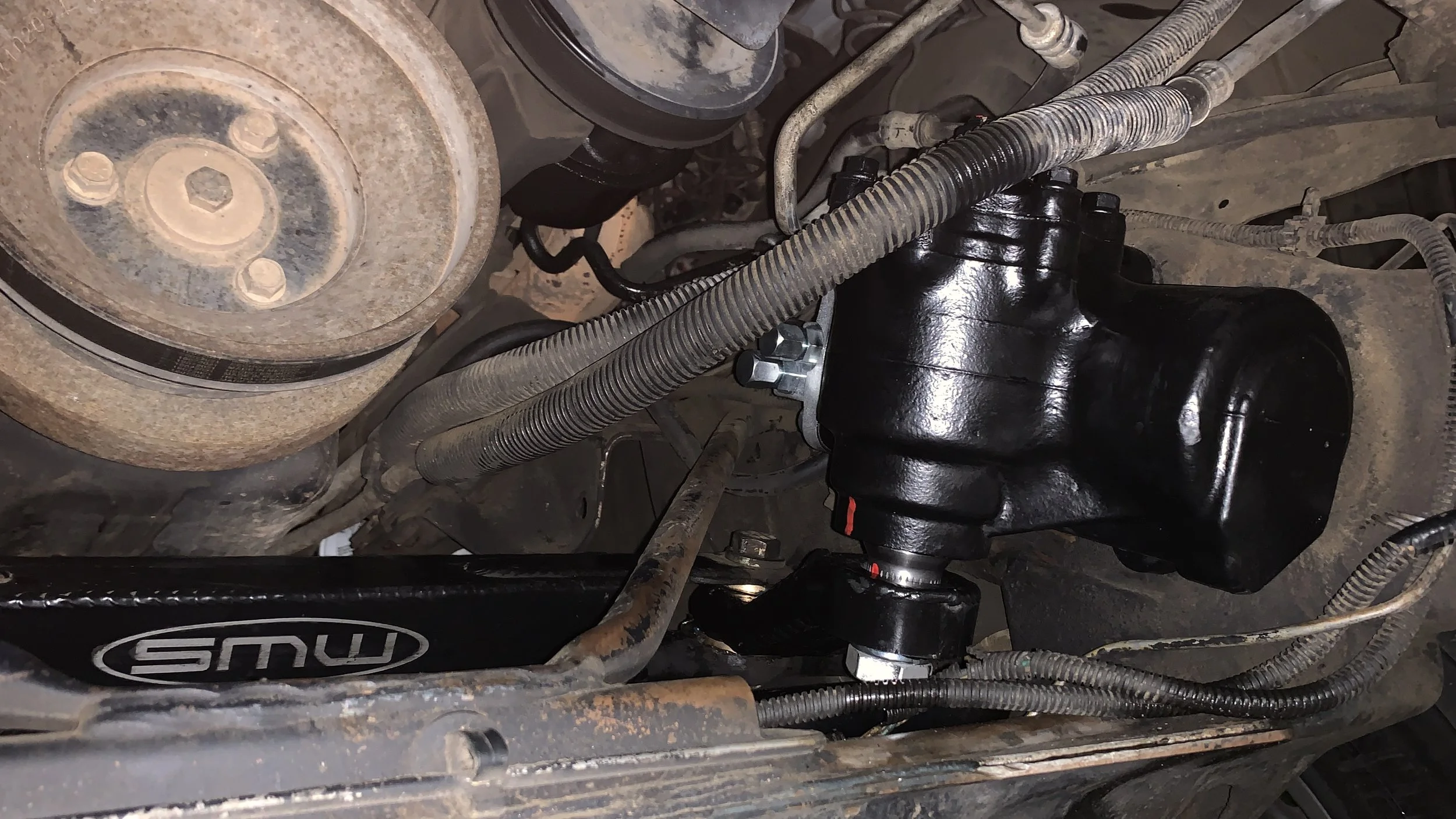 Close-up of a vehicle's engine bay showing rusted pulley, black painted component, and various hoses and wiring.
