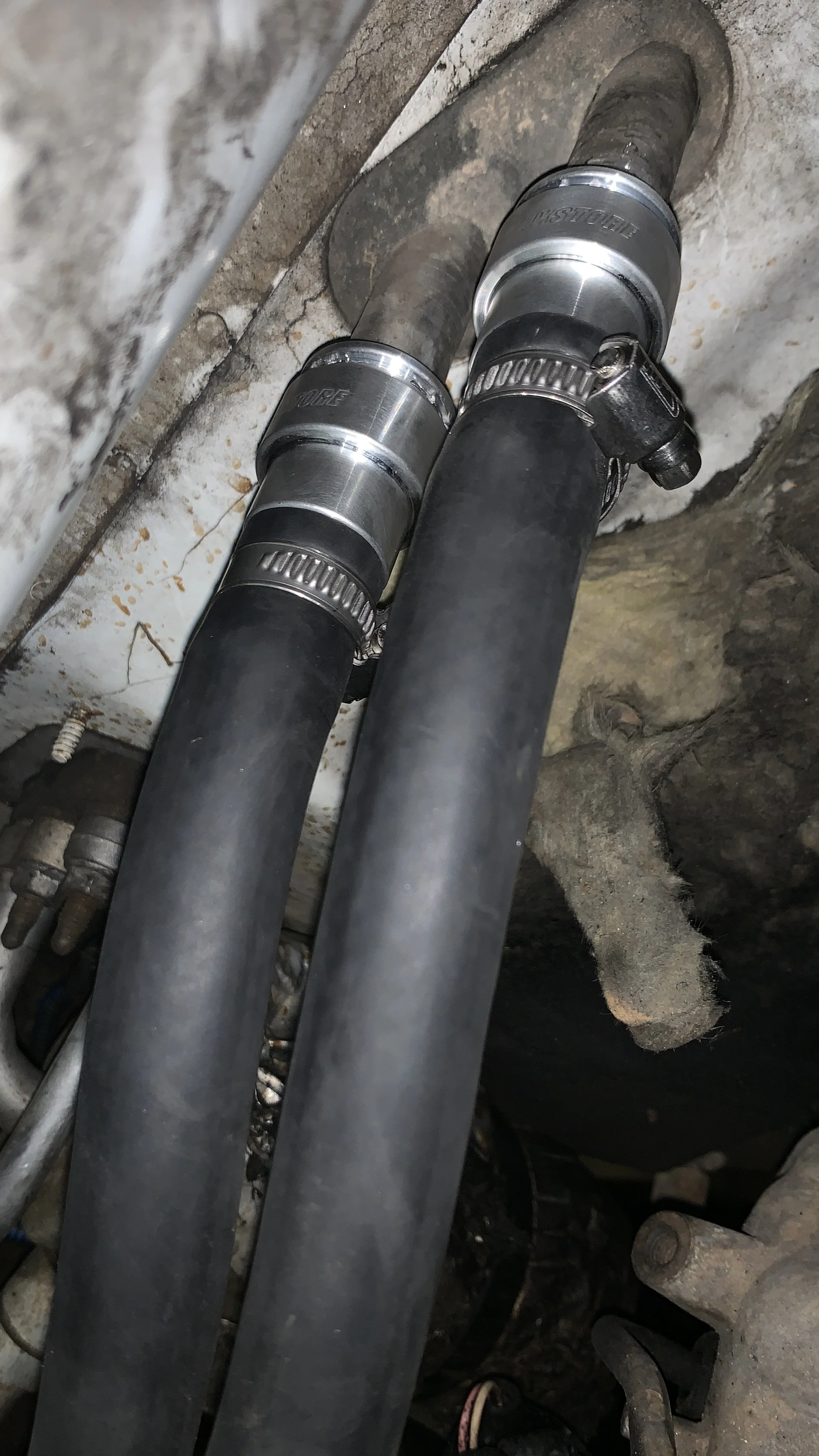 Close-up of two large, black rubber hoses with metal clamps attached, installed in a mechanical setting with a dirty wall and rocks in the background.