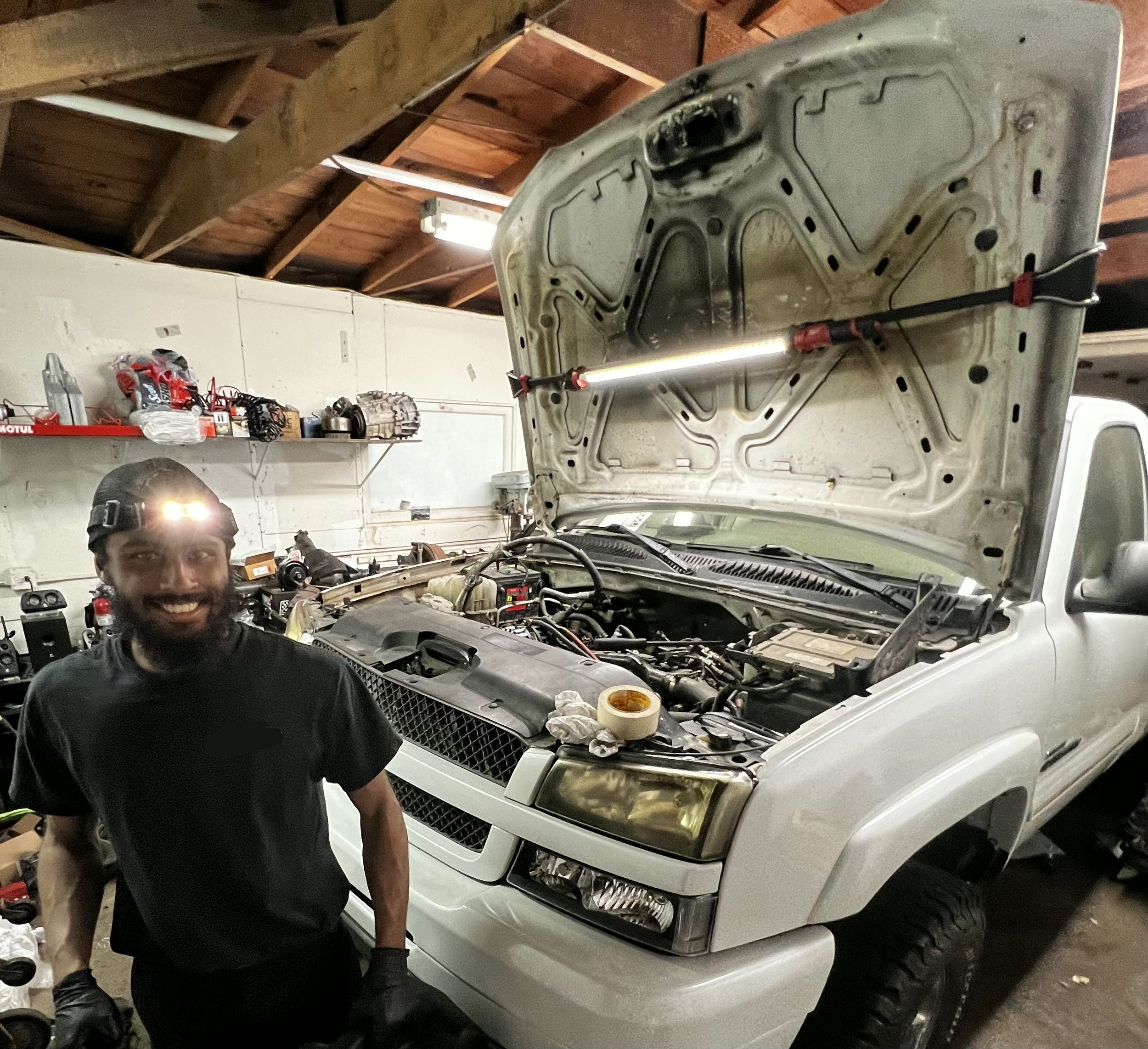 A person smiling in a workshop stands next to a white vehicle with its hood open, revealing the engine. The workshop has shelves with tools and car parts, and the person is wearing a headlamp, gloves, and dark clothing.