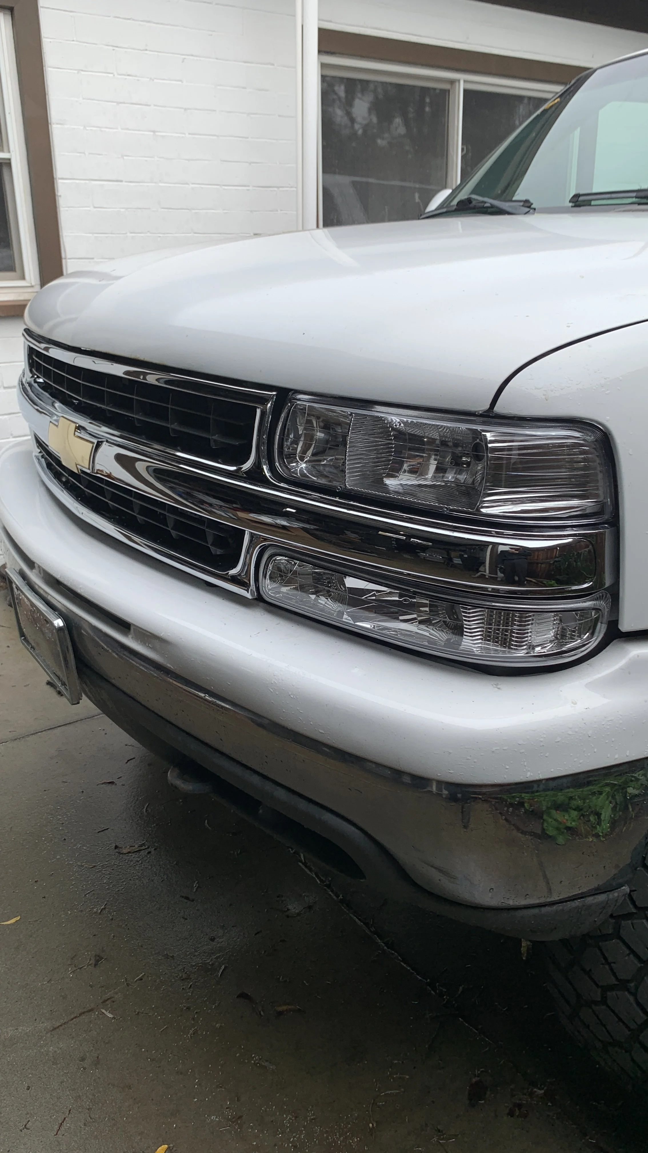 Close-up of the front of a white Chevrolet SUV, showing the grille, headlights, bumper, and part of the hood, parked near a white brick building with a window.
