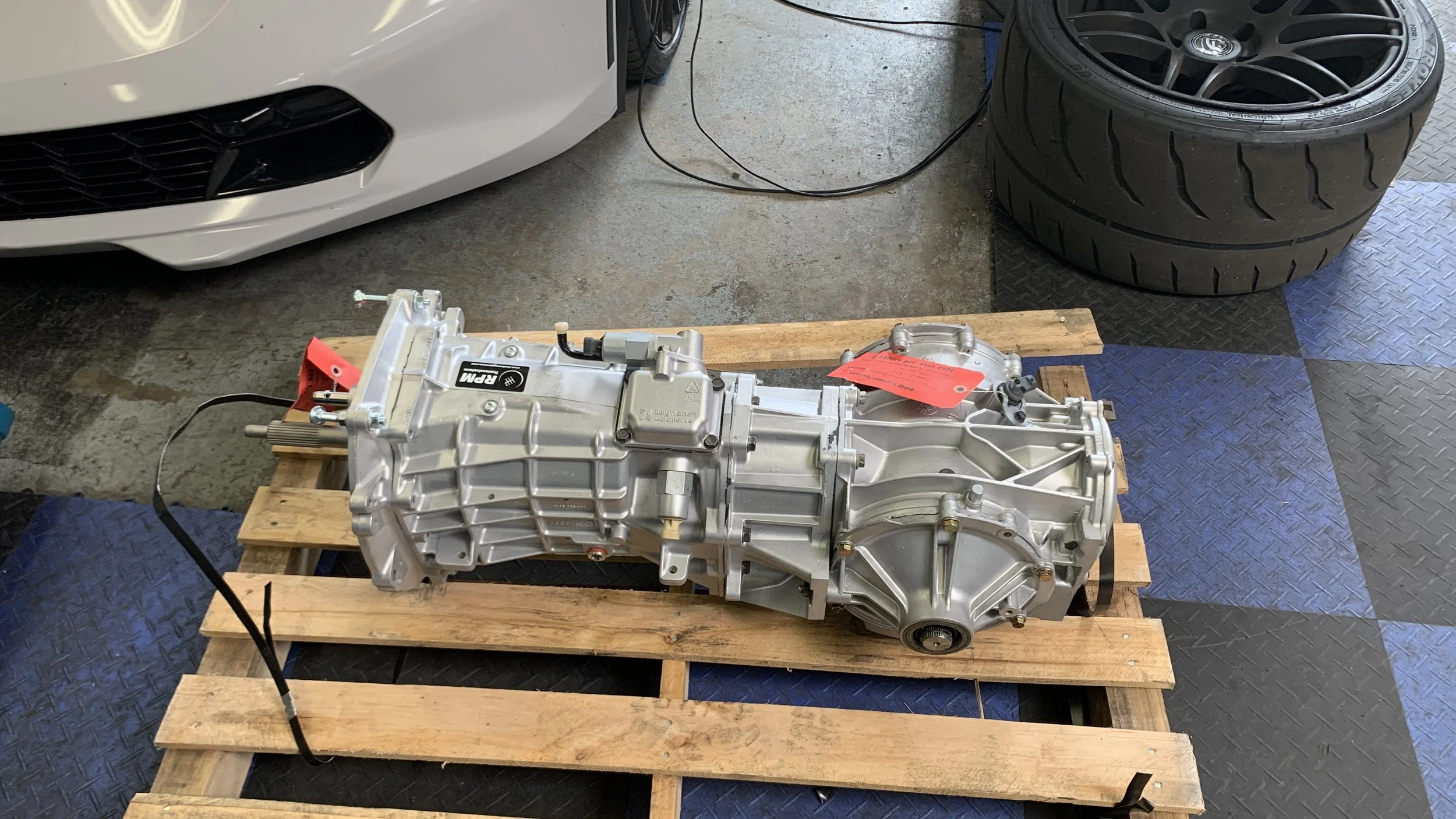 Automotive transmission on a wooden pallet in a garage next to car tires and a white vehicle