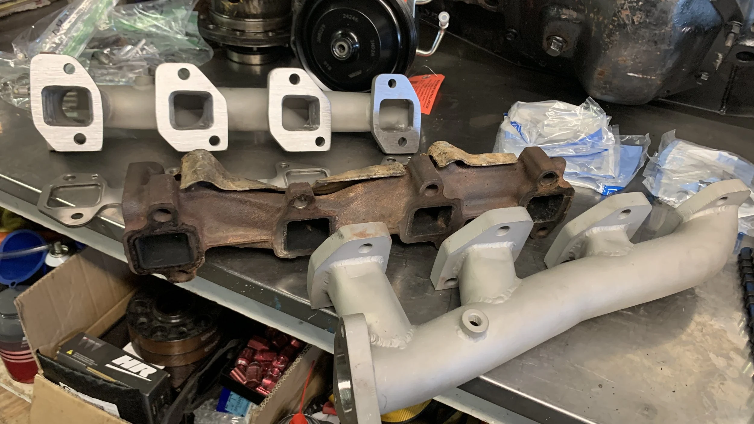 Disassembled automotive parts including a rusty exhaust manifold, a white intake manifold, a set of metal gaskets, a pulley, and some small hardware on a workbench with tools and packaging materials.