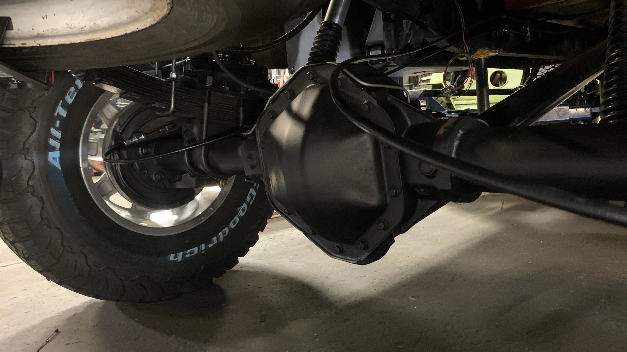 Close-up view of a vehicle's drivetrain and rear suspension components, including a tire with the 'All-Terrain' logo, differential, and suspension springs.