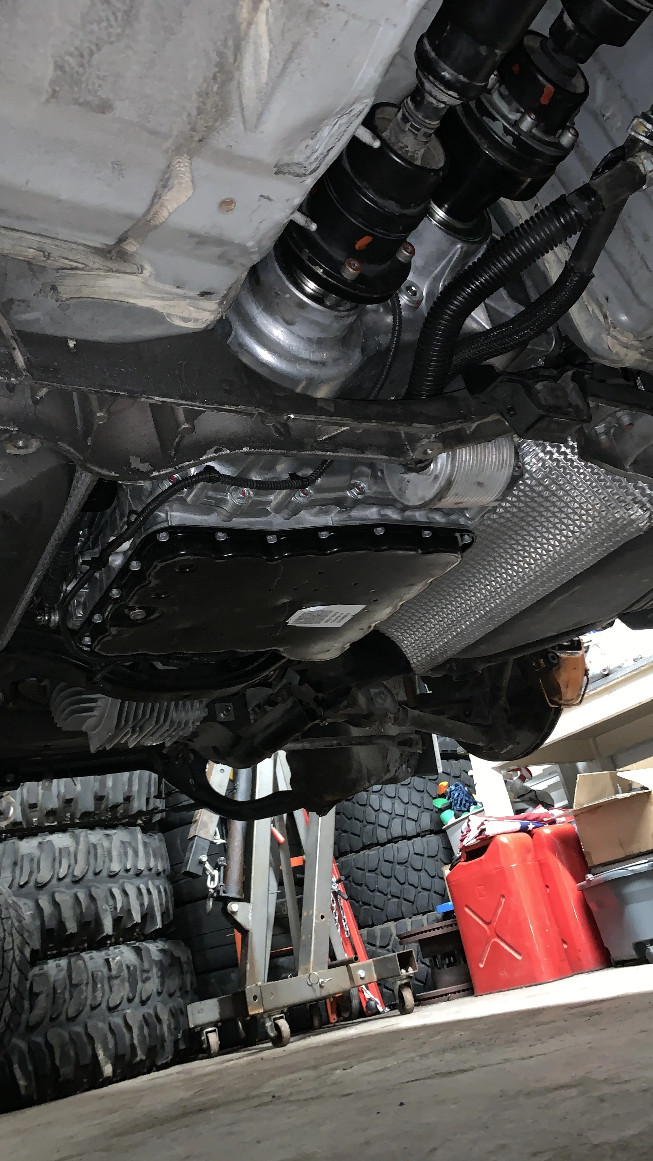 Underneath view of a car with visible suspension, oil pan, and exhaust heat shield, in a garage with tools, tires, and a red fuel container in the background.
