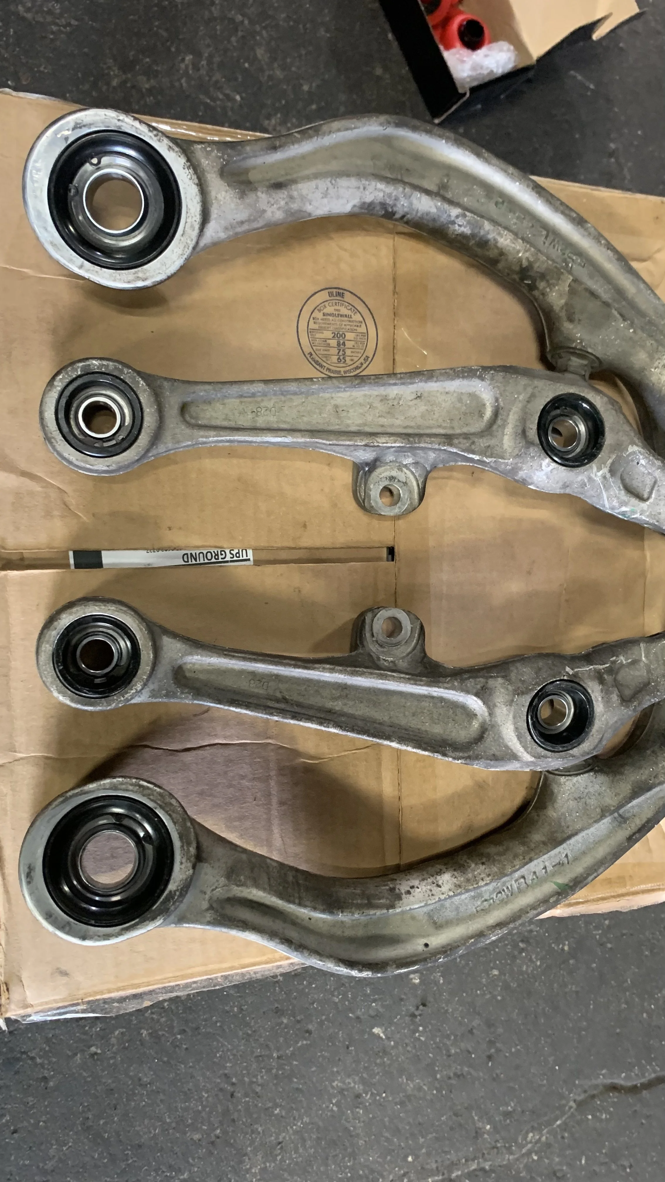 Four metal control arms with ball joints attached, used in a vehicle's suspension system, laid on a cardboard surface.