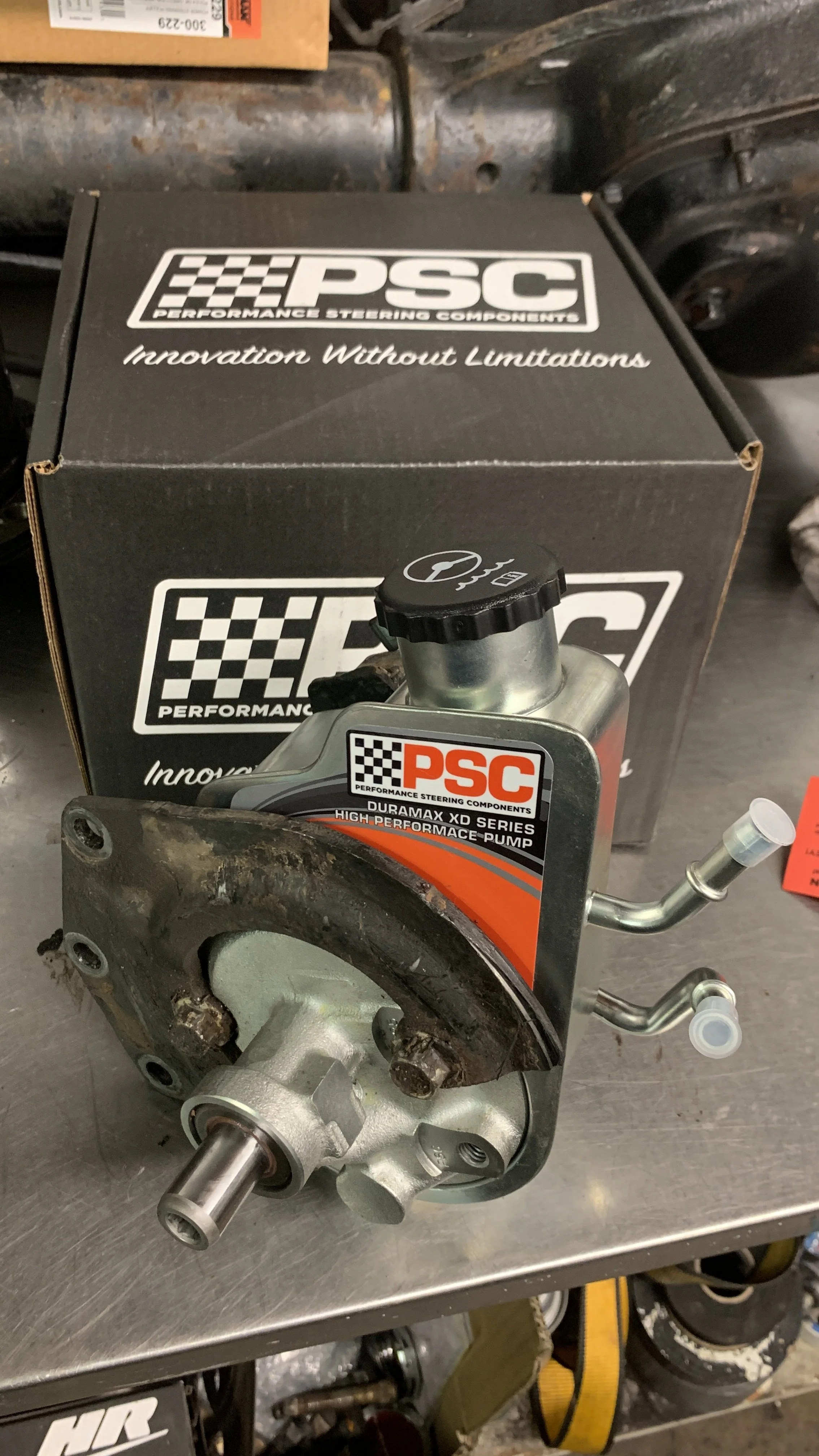 A PSC high-performance pump with attached metal fittings, placed on a workbench in front of a black PSC Performance Steering Components box.