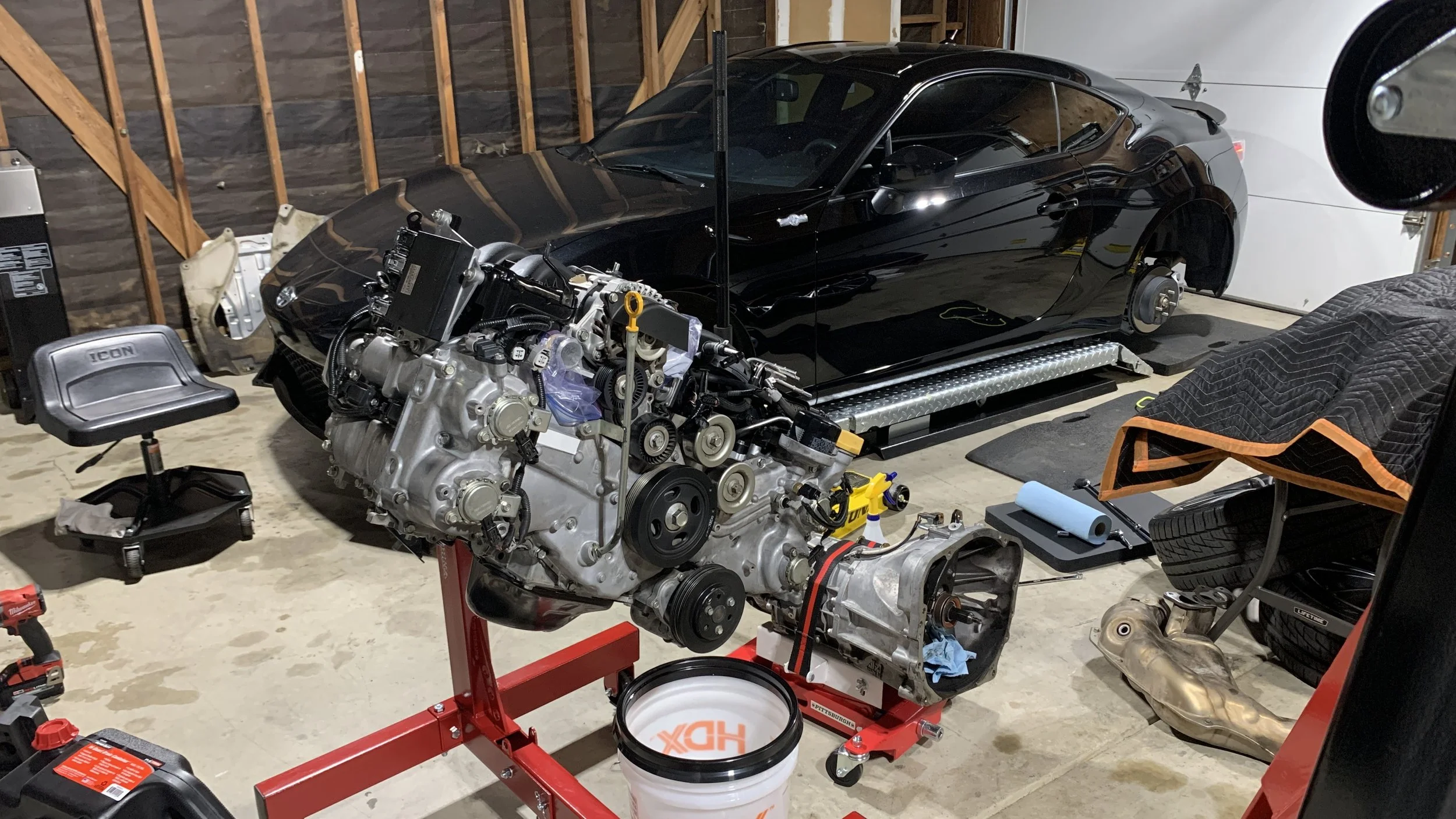 A race car engine mounted on a red engine stand in a garage, with a black sports car body without wheels visible in the background, and garage tools and supplies scattered around.