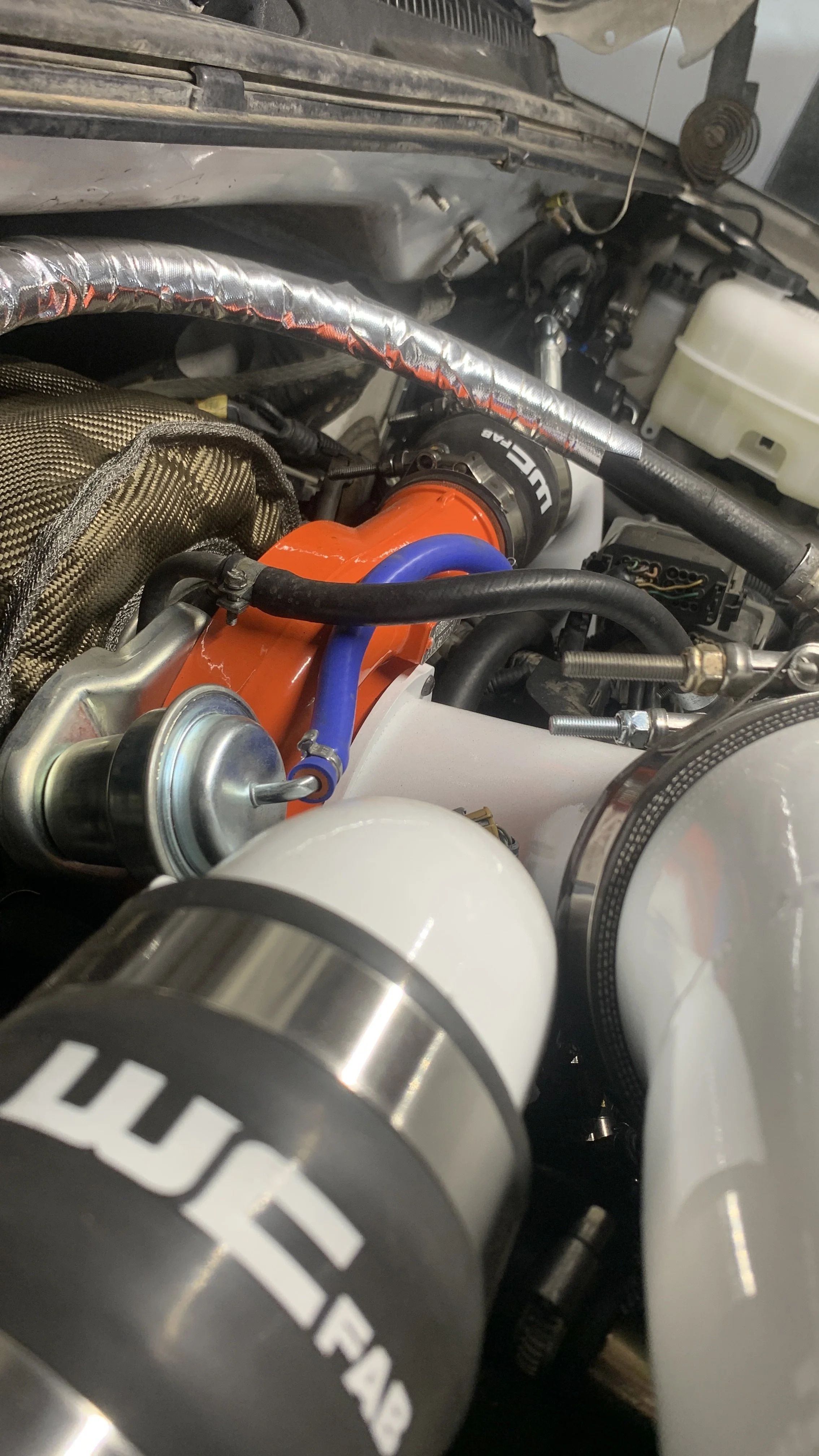 Close-up of a car engine showing various components including a white canister, a metallic part with a black label, hoses, and an orange part, with some wiring and a matrix metal mesh cover.