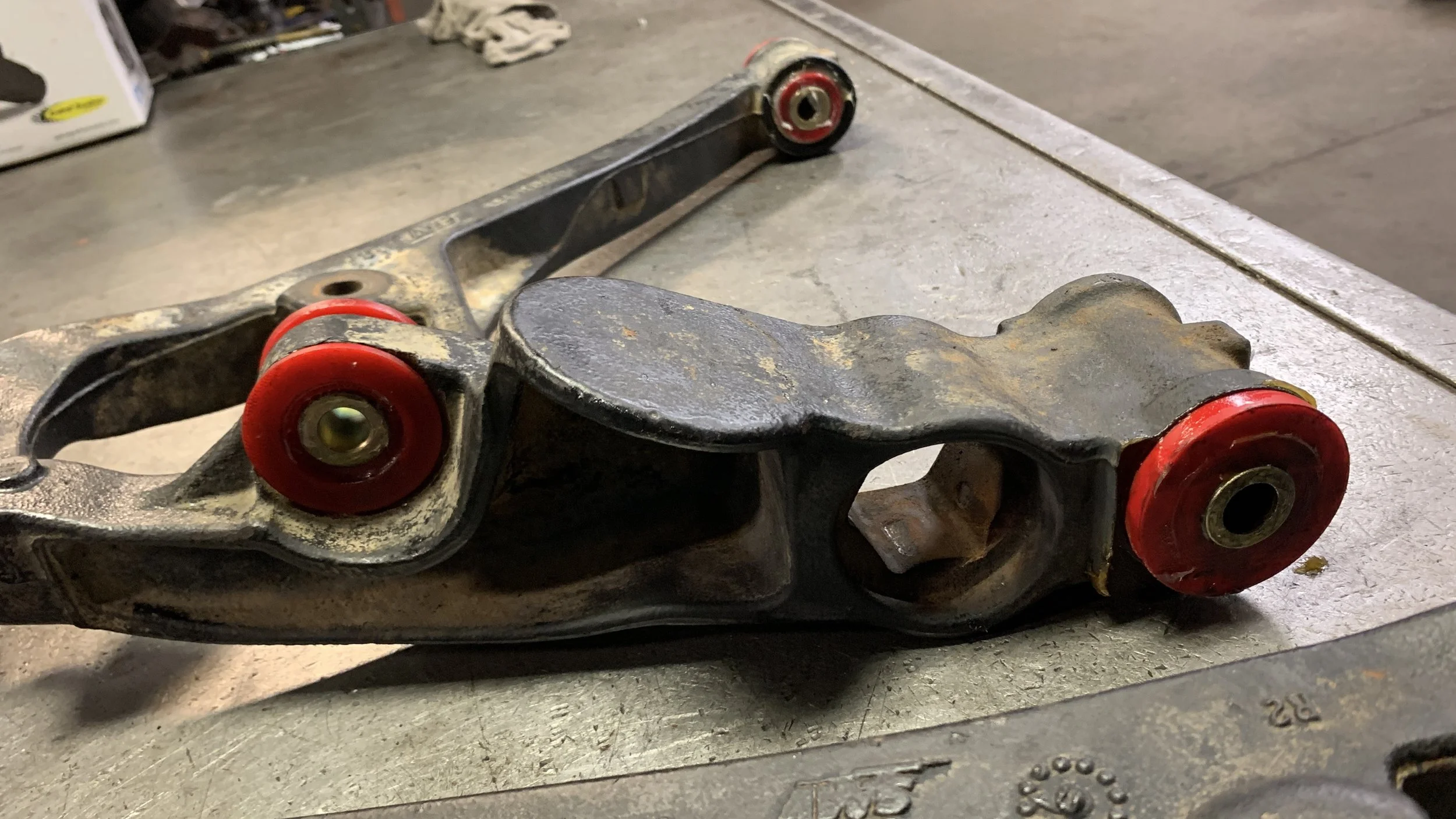 Automotive suspension control arm with mounted red bushings, resting on a metal work surface.