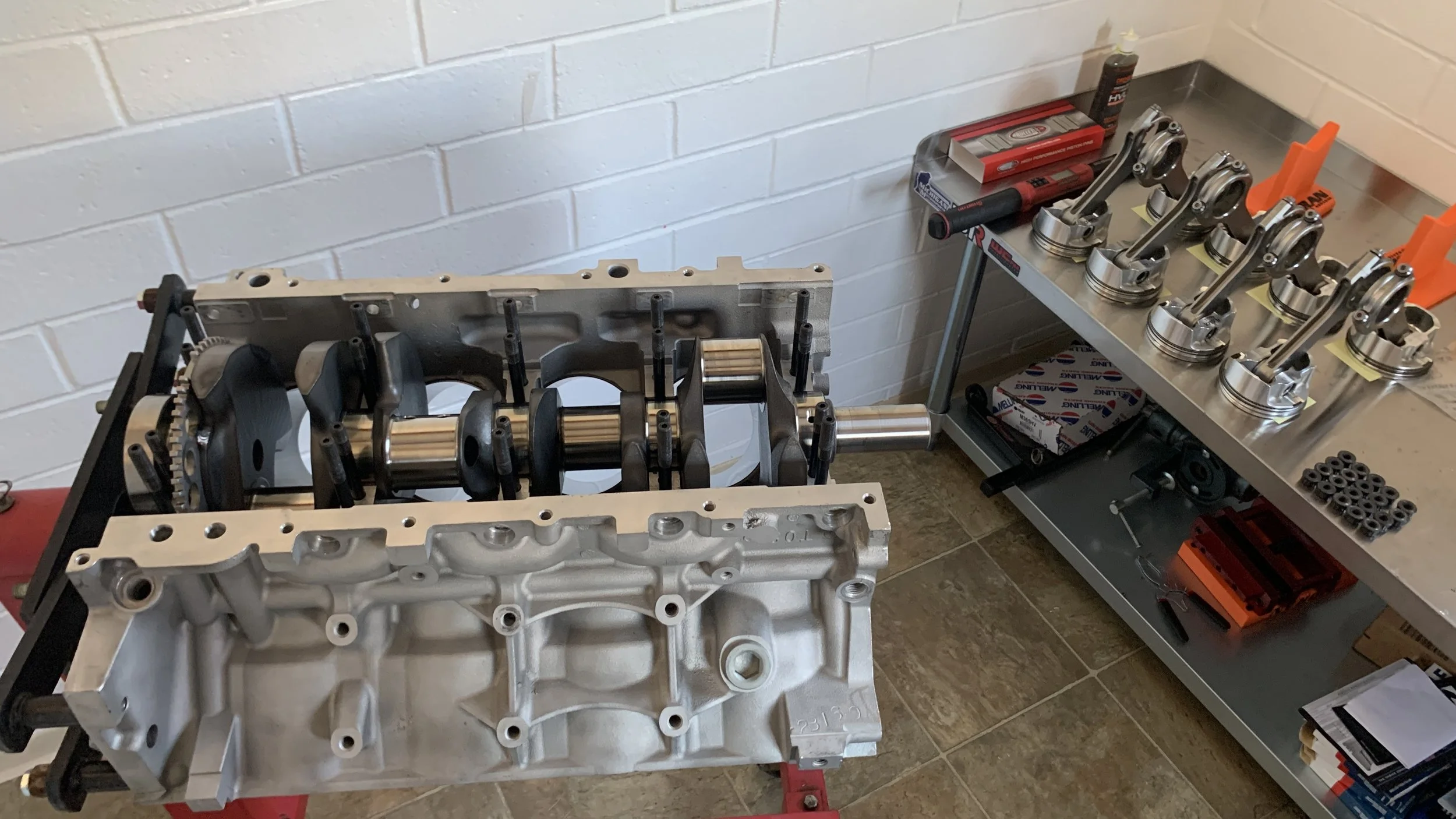 Automotive engine parts, including a disassembled engine block and pistons, on workbenches in a workshop with white brick walls.