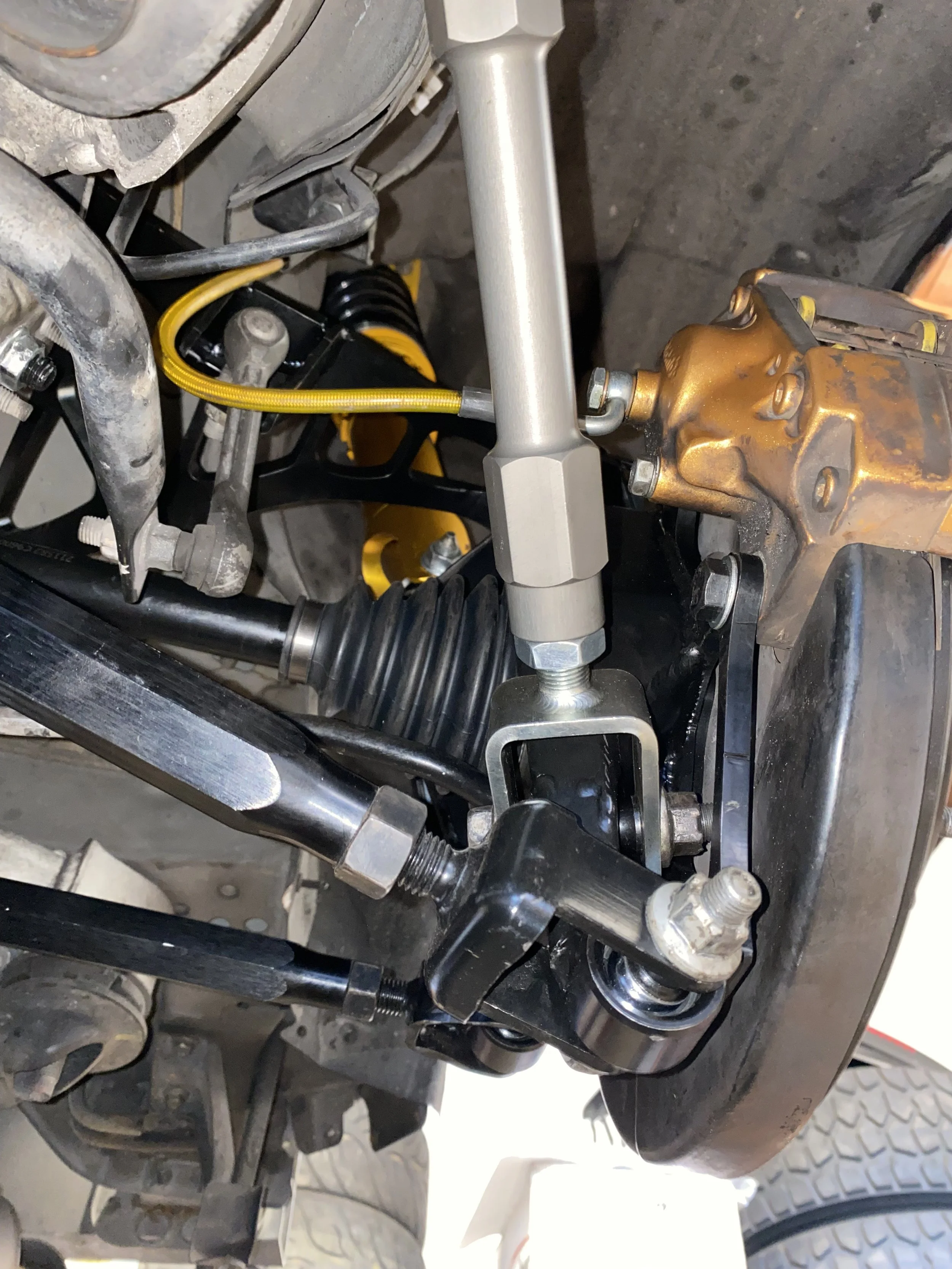Close-up of a vehicle's suspension and steering components, including a Gothic-style steering rack, a yellow coilover shock, and various metal linkages.