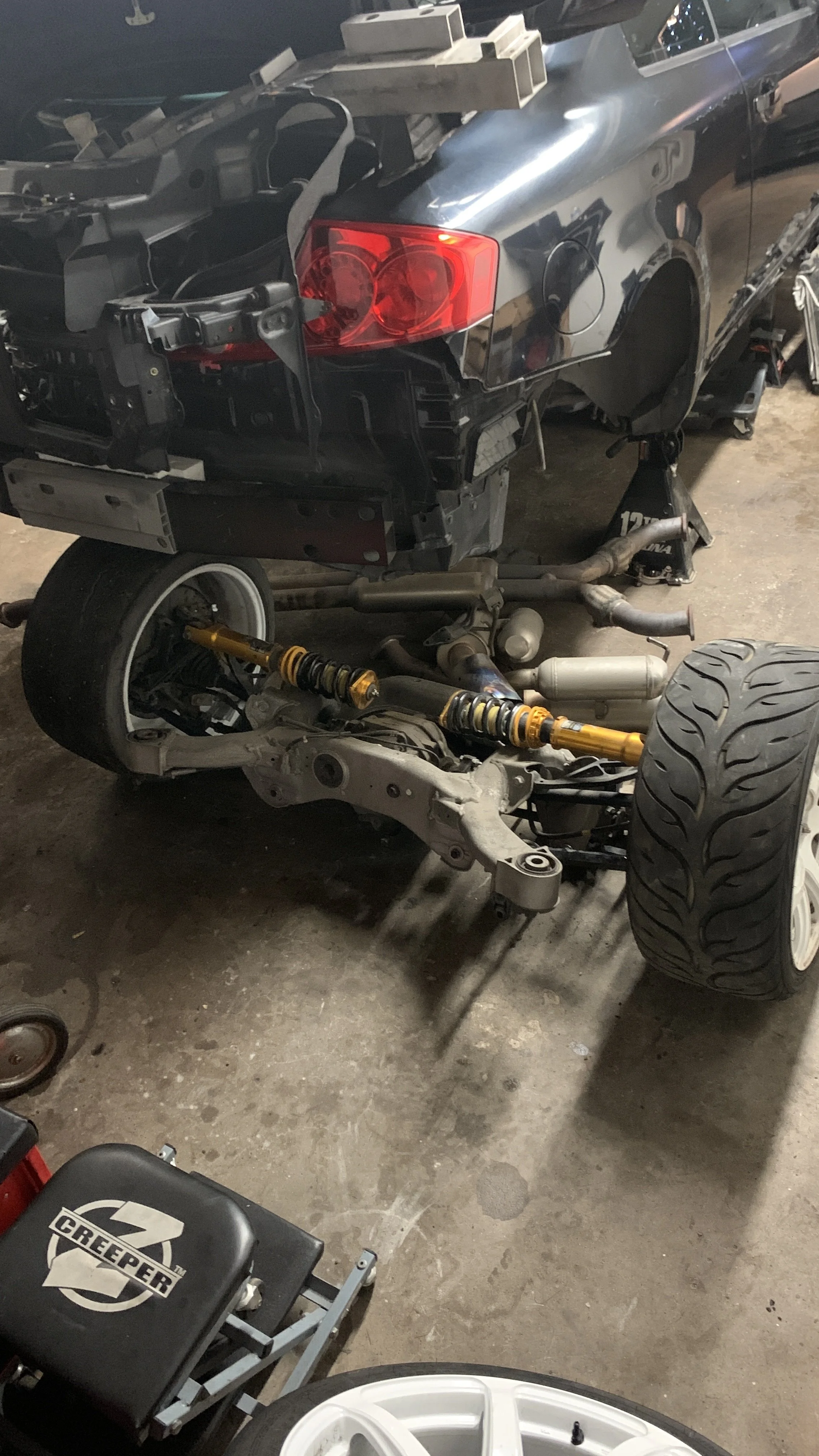 A partially disassembled black sports car is on a lift in a garage, with the rear bumper removed revealing internal components. The rear suspension and exhaust system are visible, and the car is supported by jack stands. A wheel with a white rim is o