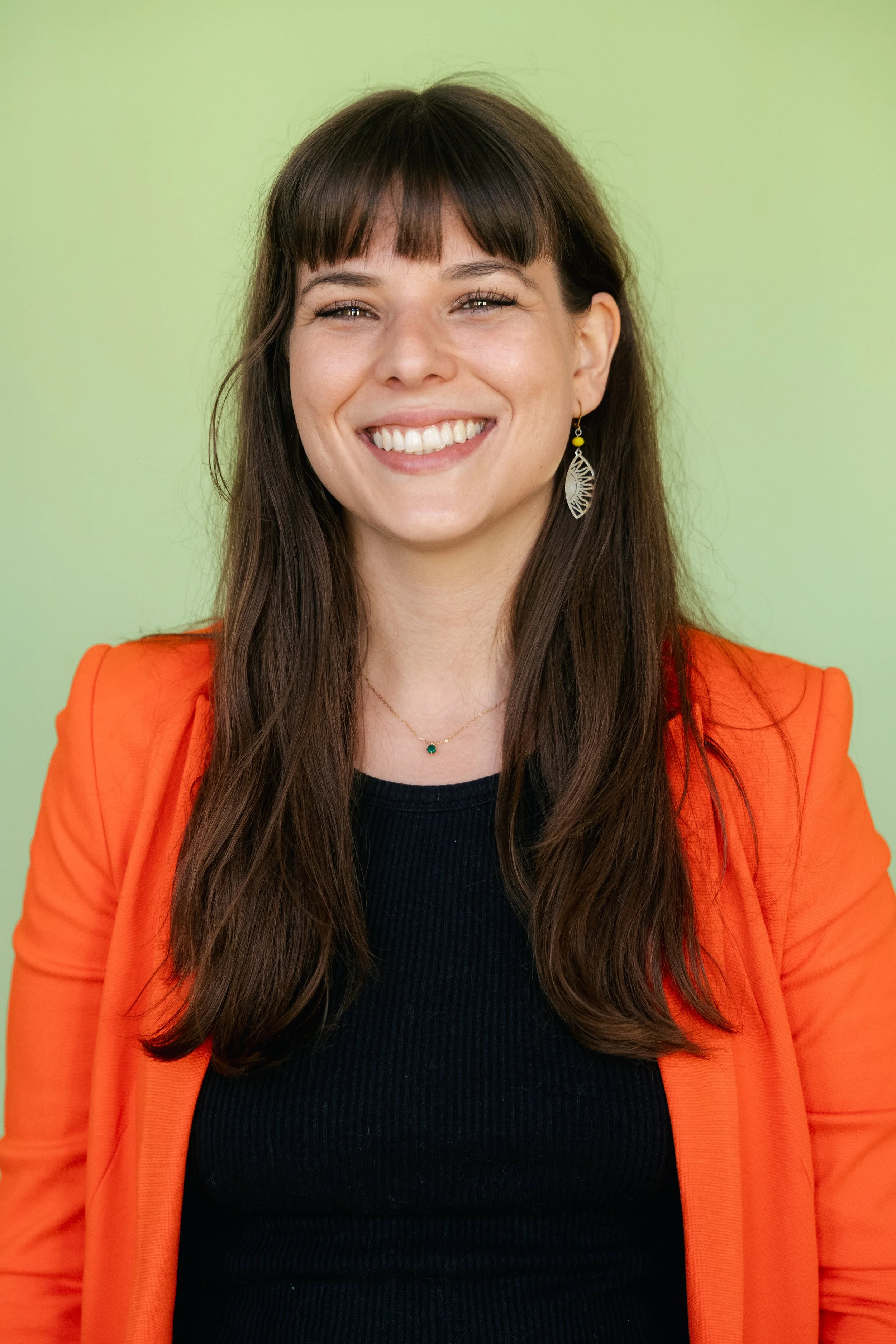 A woman with long brown hair, wearing an orange blazer, black top, and earrings, smiling against a light green background.