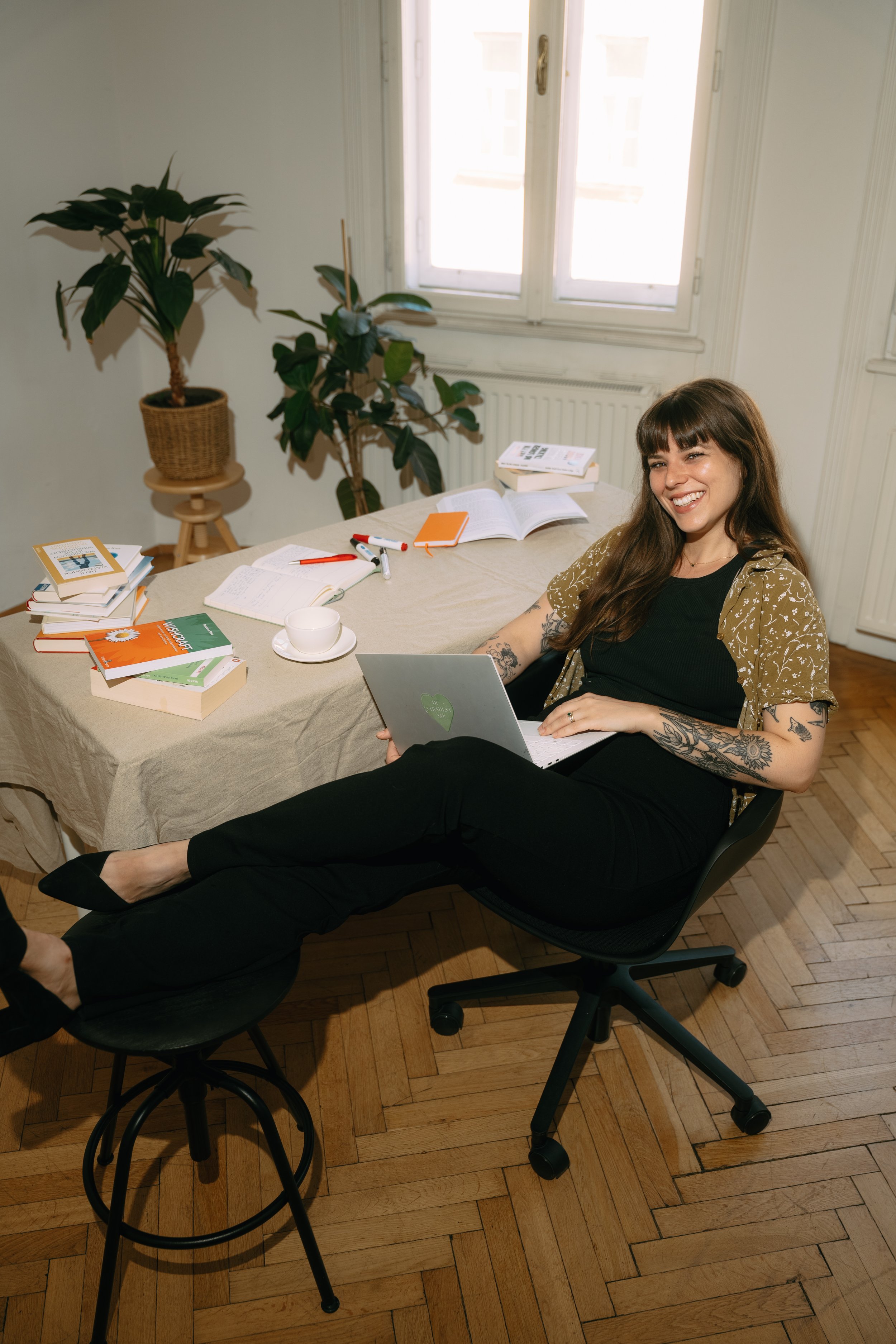A woman with long brown hair and tattoos on her arms, sitting in a black office chair, smiling and reading a notebook. She is in a room with a large window, a table with books, markers, an open notebook, and a cup. There is a potted plant on a stand beside the window and a wooden floor.