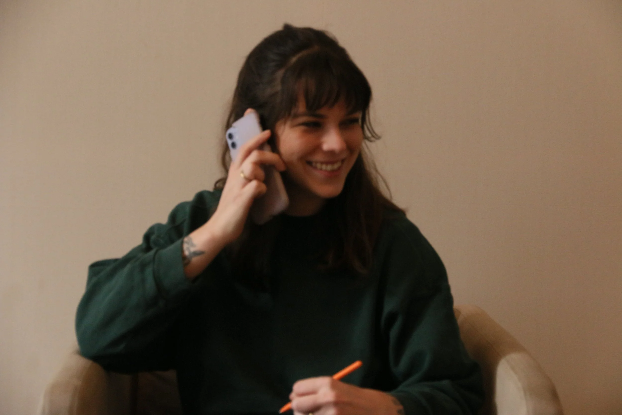 A woman with dark brown hair tied in a ponytail with bangs, smiling, talking on a mobile phone, wearing a dark green jacket, sitting at a table with an orange pen in her hand.