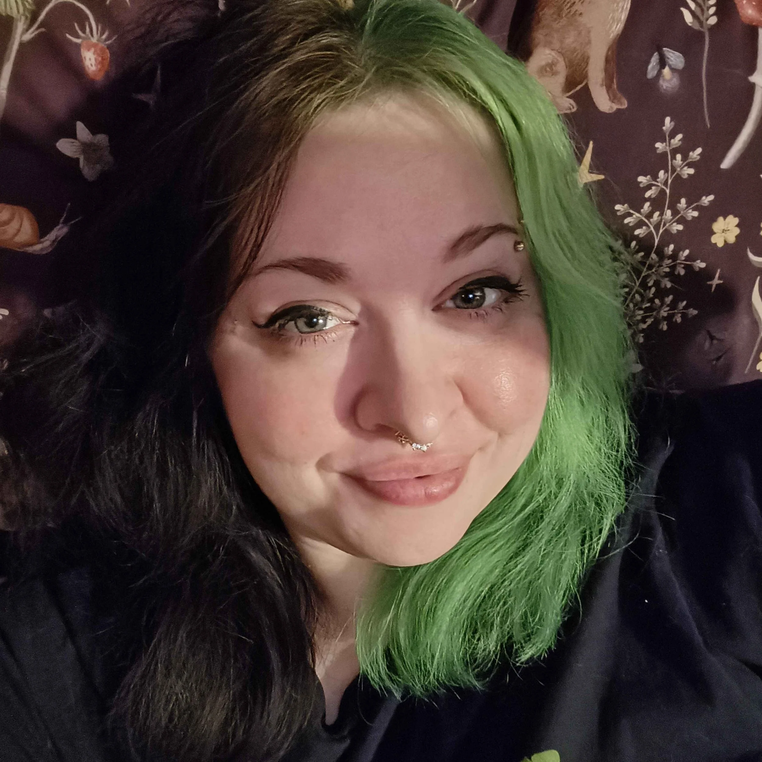 Li smiles at the camera, they have half black and half green hair and piercings.