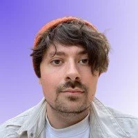 Joe smiles at the camera. He has short brown hair and an orange hat.