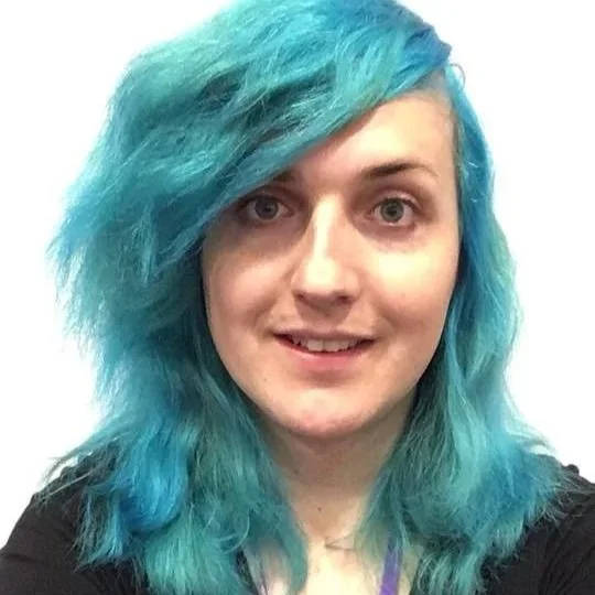 Laura smiles at the camera, wearing a black top. She has bright blue hair.