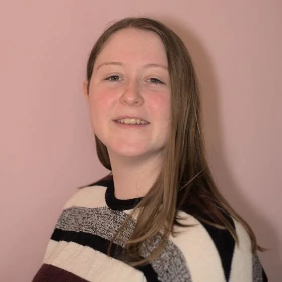 Megan smiles at the camera. They have long brown hair and are wearing a striped top.