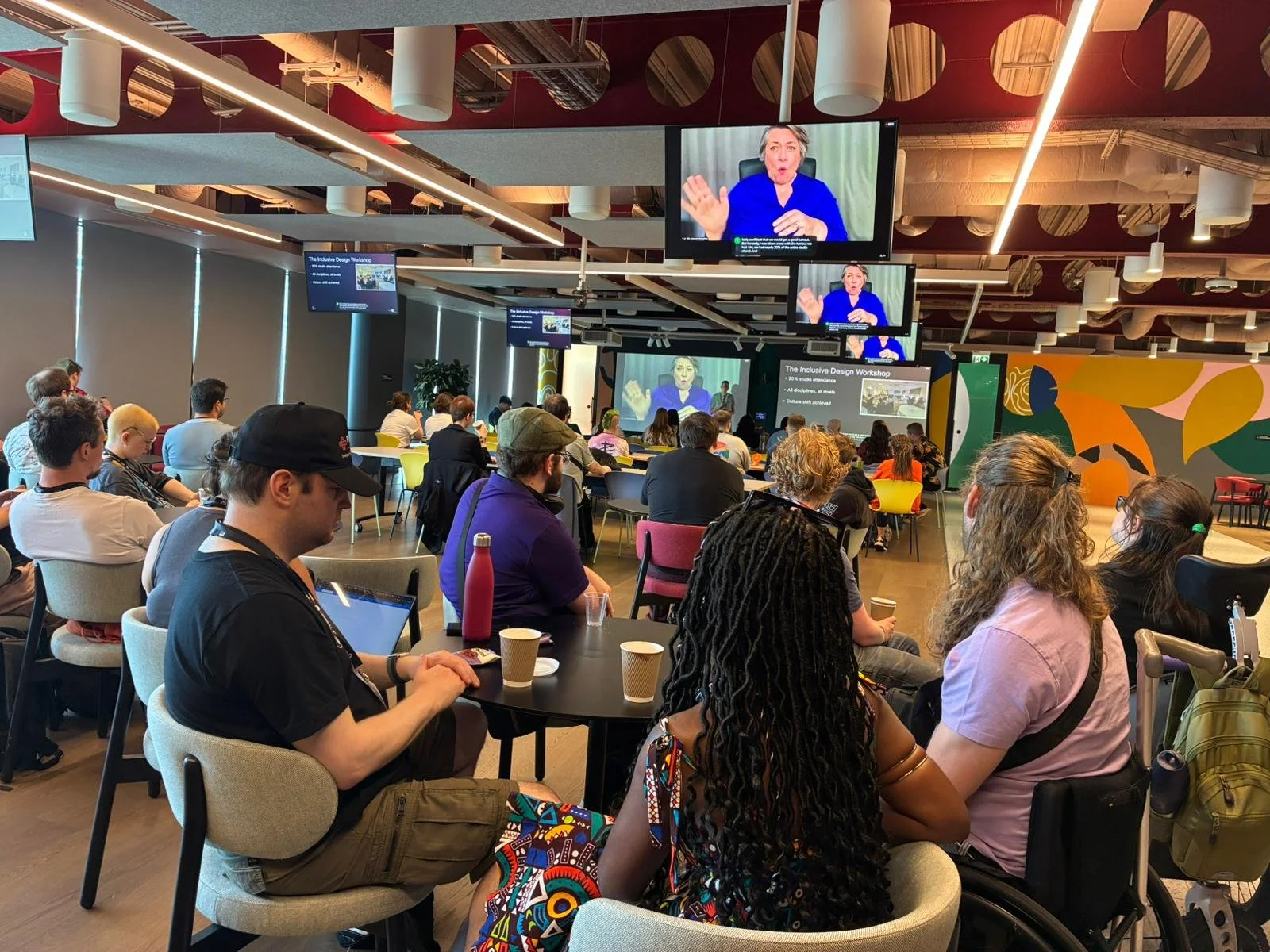 A photo of the event, many attendees are sat at round tables. Screens on the ceiling and at the back of the event show slides and BSL interpretation.