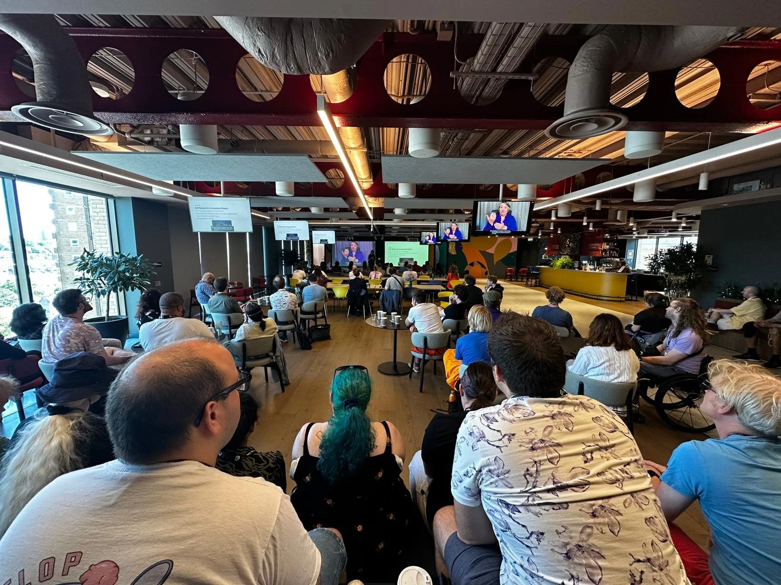A photo of the gathering in person in the Unity offices. Many people sit at small tables, desks and bleacher seating. Two large screens at the back of the room show talk slides and BSL interpreter. Small TVs on the ceiling mirror the big screens.