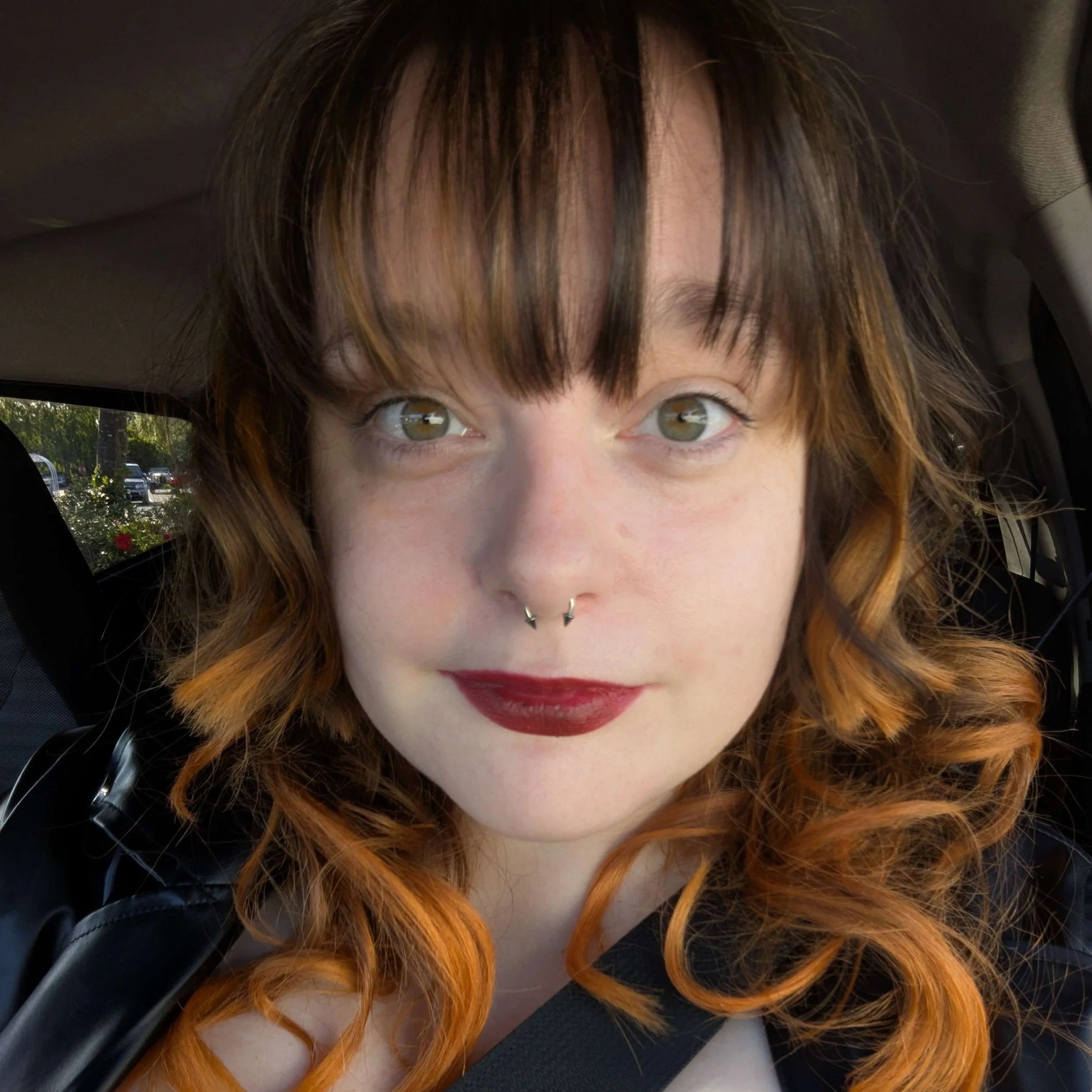 Steph smiles at the camera. They have dark hair with orange highlights and are wearing deep red lipstick.