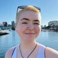Chloe smiles at the camera, standing in front of a waterfront. They have shaved shirt hair.