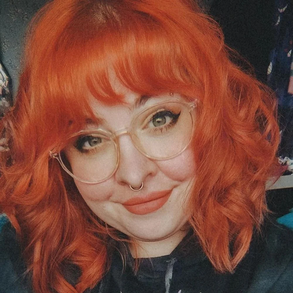 Li smiles at the camera. They have bright orange hair, matching lipstick and clear-frames glasses.