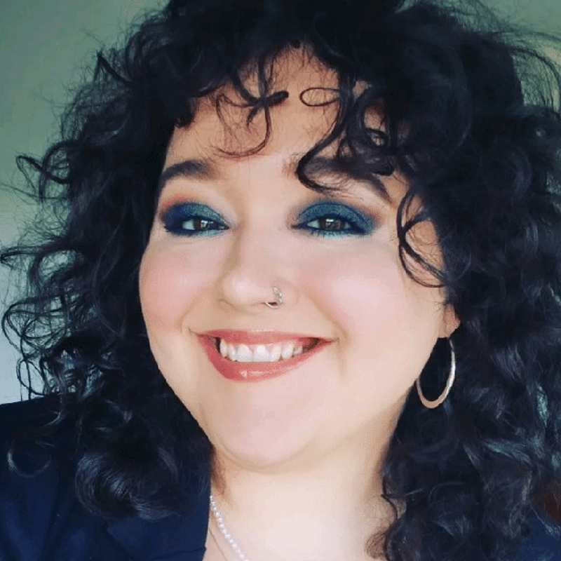 Ameliane smiles at the camera. She had dark curly hair and dark blue eyeshadow.