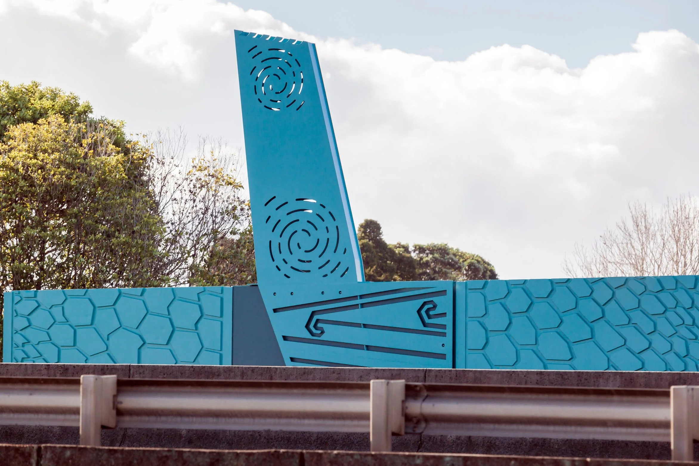 A blue decorative panel with geometric patterns, circular cutouts, and line art, mounted on a structure, with trees and a partly cloudy sky in the background.