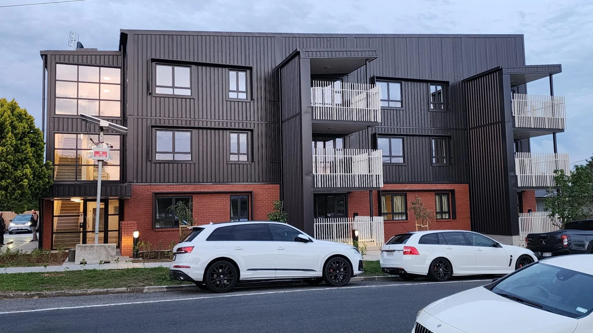 A modern multi-story apartment building with a brick base and dark metal siding, with white balconies on each level, and parked cars along the street in front.