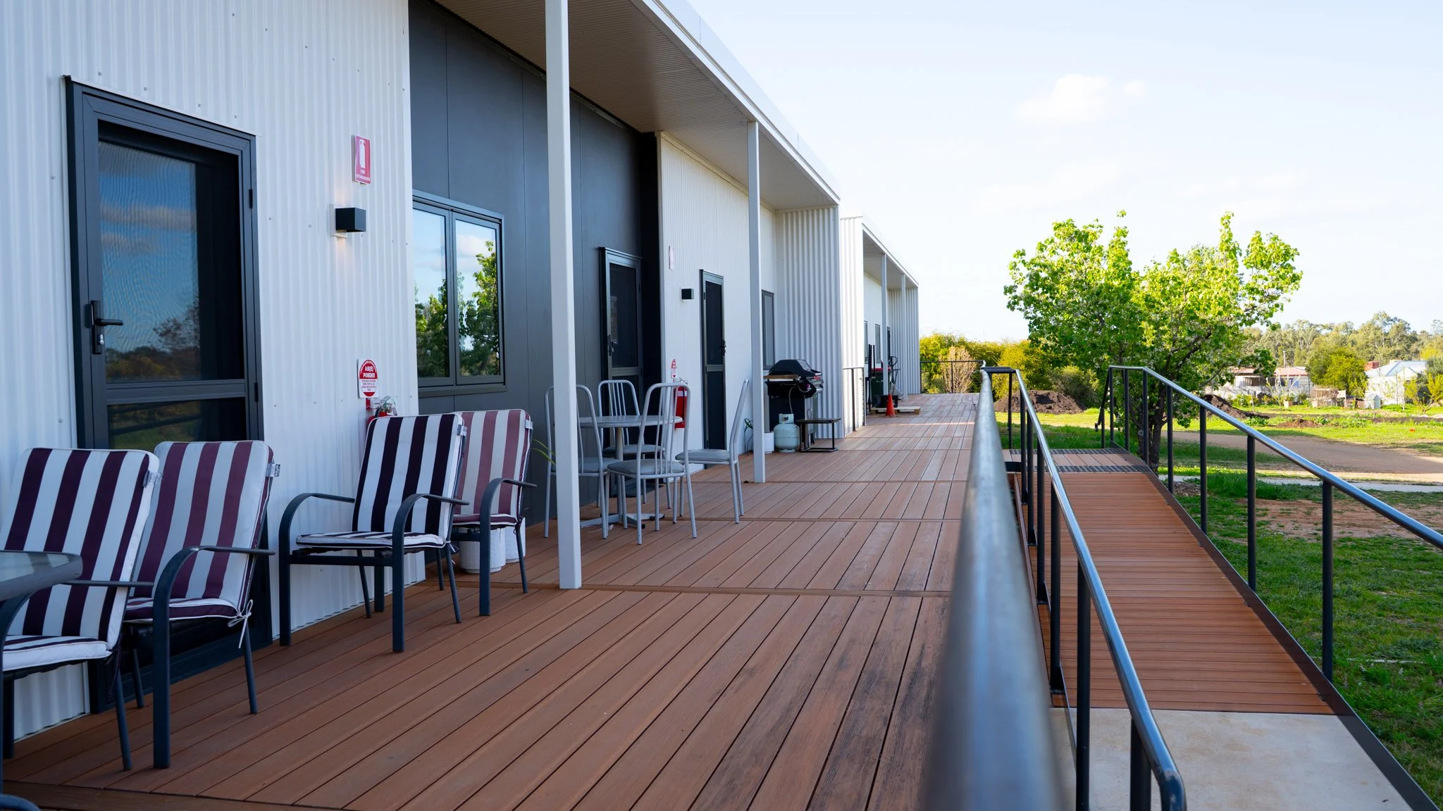 A wide outdoor balcony with wooden flooring, black railing, several chairs, a grill, and green trees in the background.