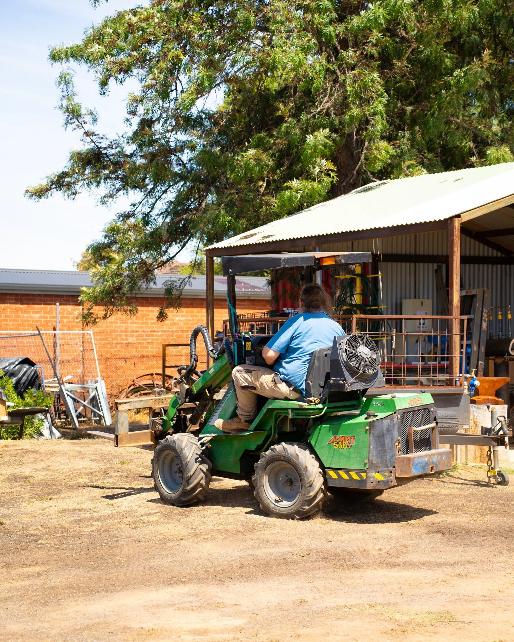 A person operating a green compact excavator on a dirt surface in a backyard with a large leafy tree and a brick building in the background.