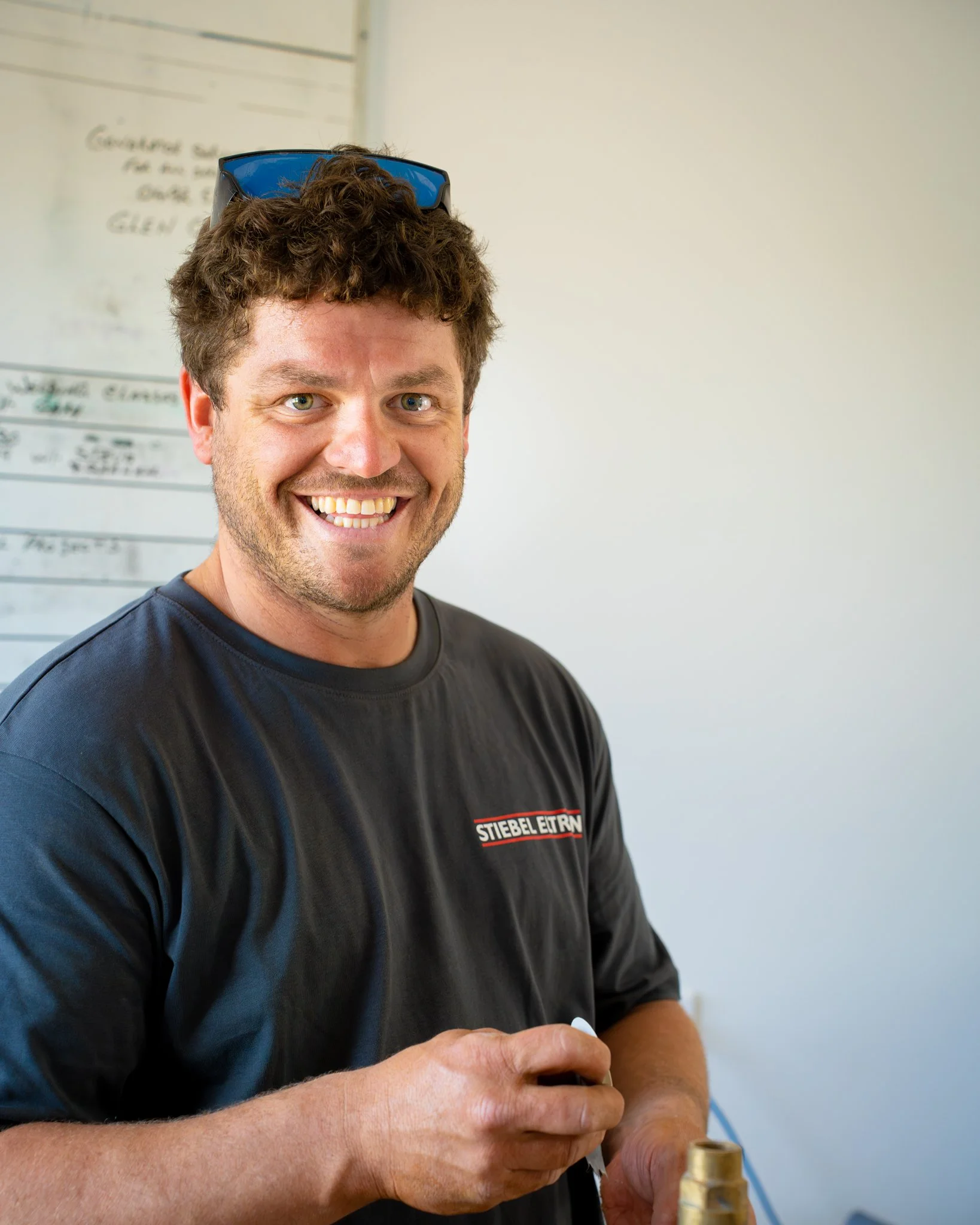 A man with curly brown hair and green eyes smiling, wearing sunglasses on his head, a black t-shirt with a logo that says 'STIEBEL ELTRON', holding a small object in his hands in front of a white wall background.